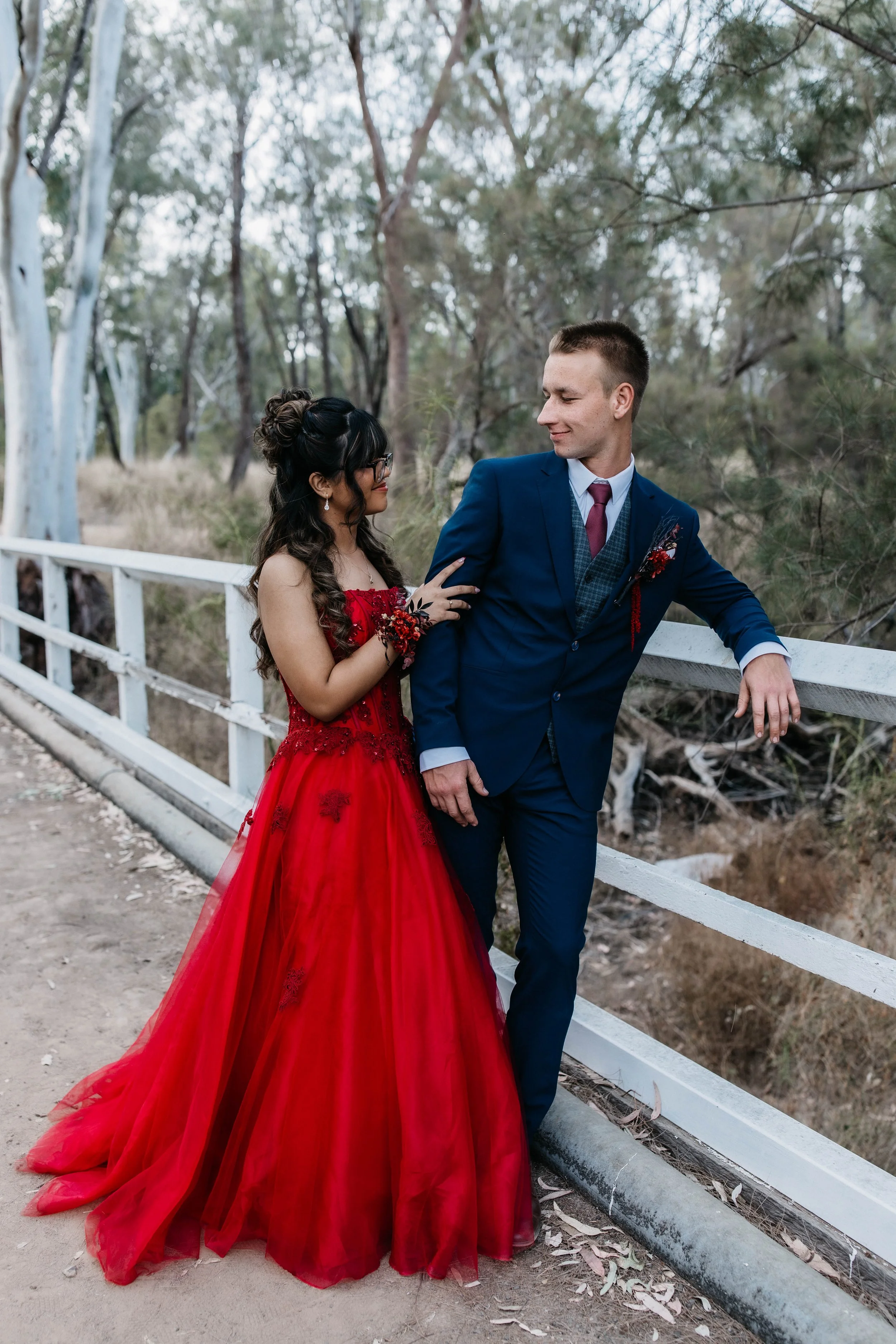 A couple dressed in formal attire standing on a bridge in a wooded area. The woman wears a red gown and the man wears a blue suit. They are looking at each other.