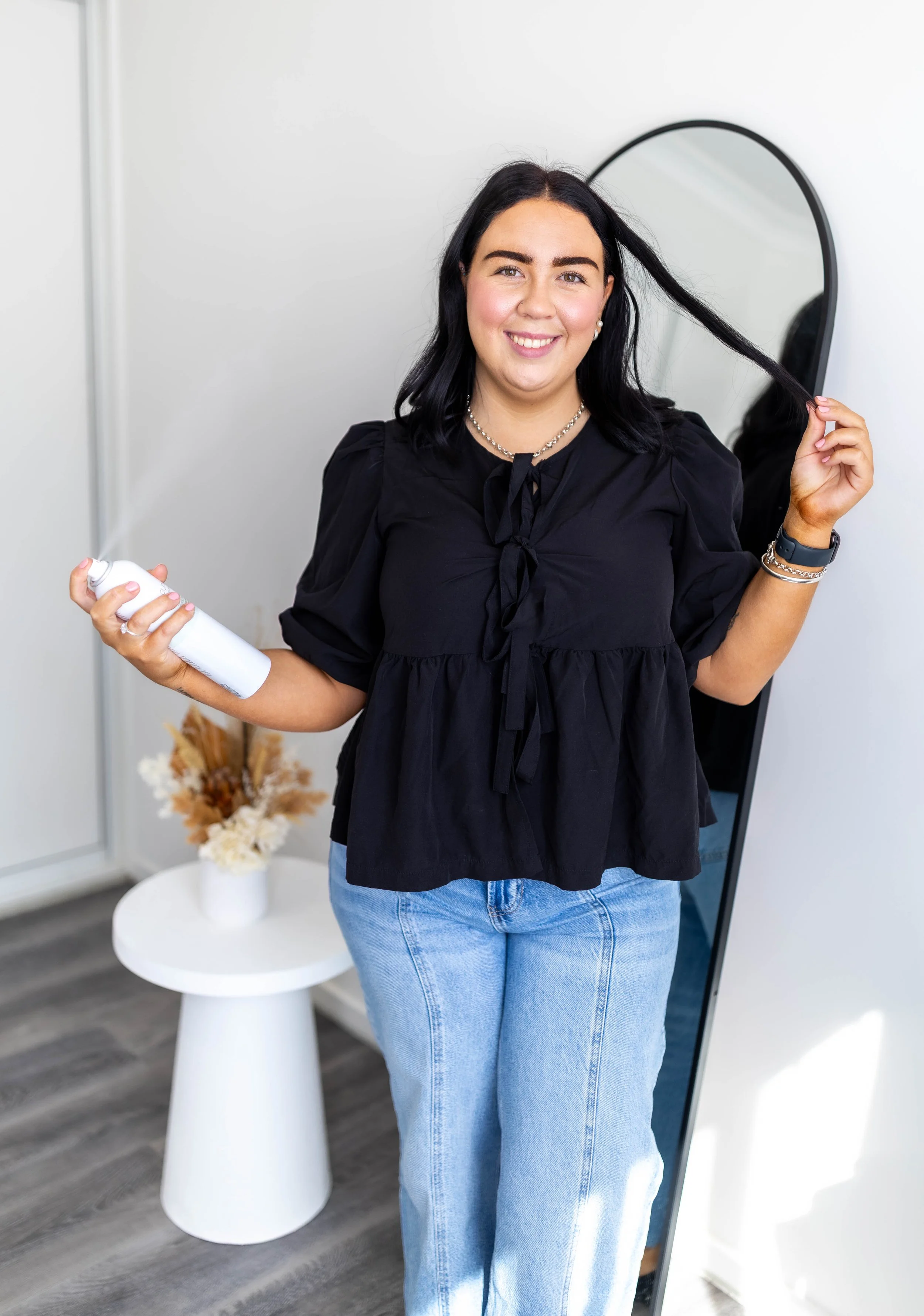 A woman with dark hair and a black shirt standing in front of a mirror, holding a bottle of hair spray in one hand and a section of her hair in the other, smiling at the camera.
