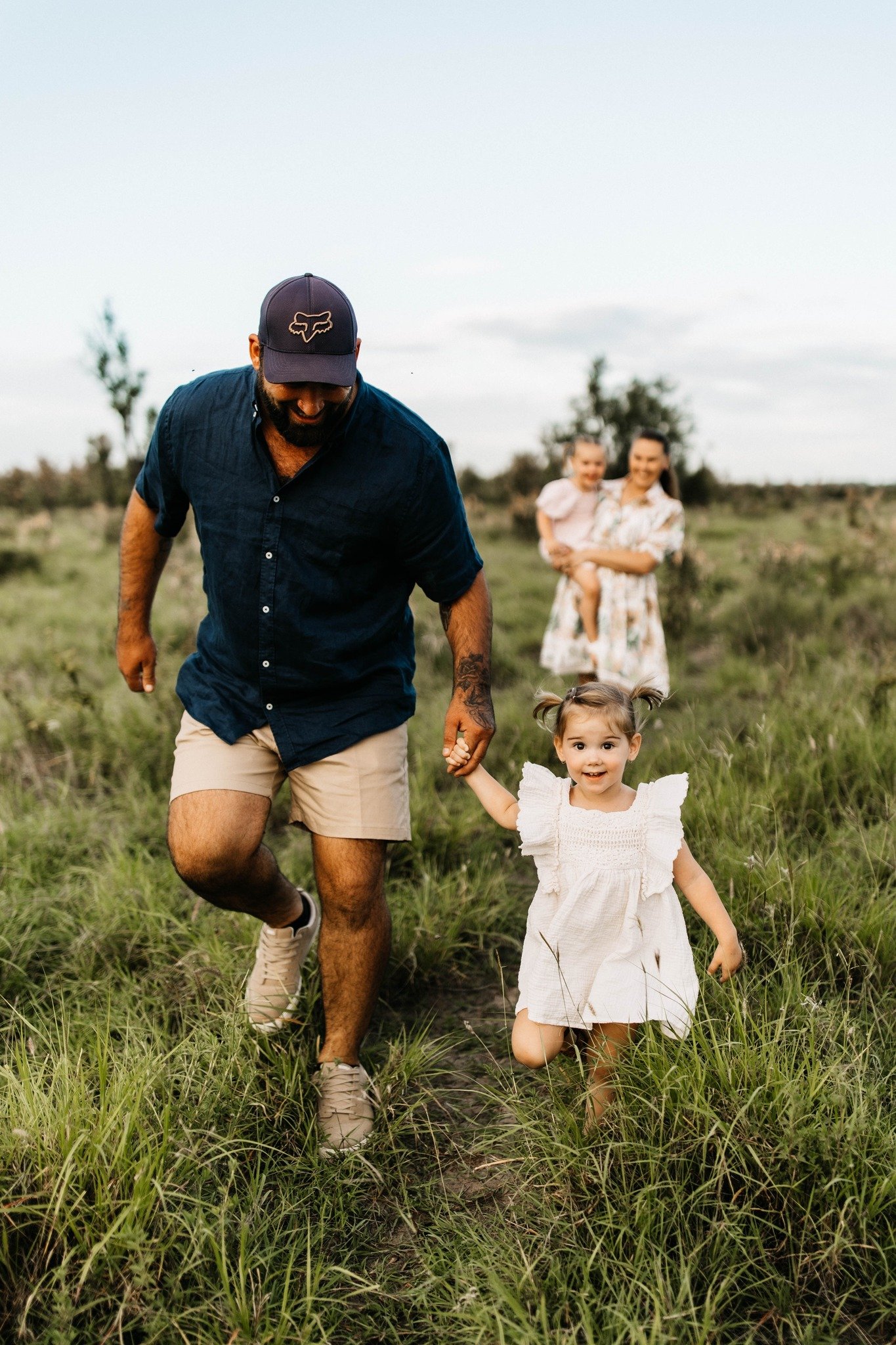 A man and a young girl holding hands and walking through a grassy field, with a woman and a young girl in the background, all outdoors on a cloudy day.