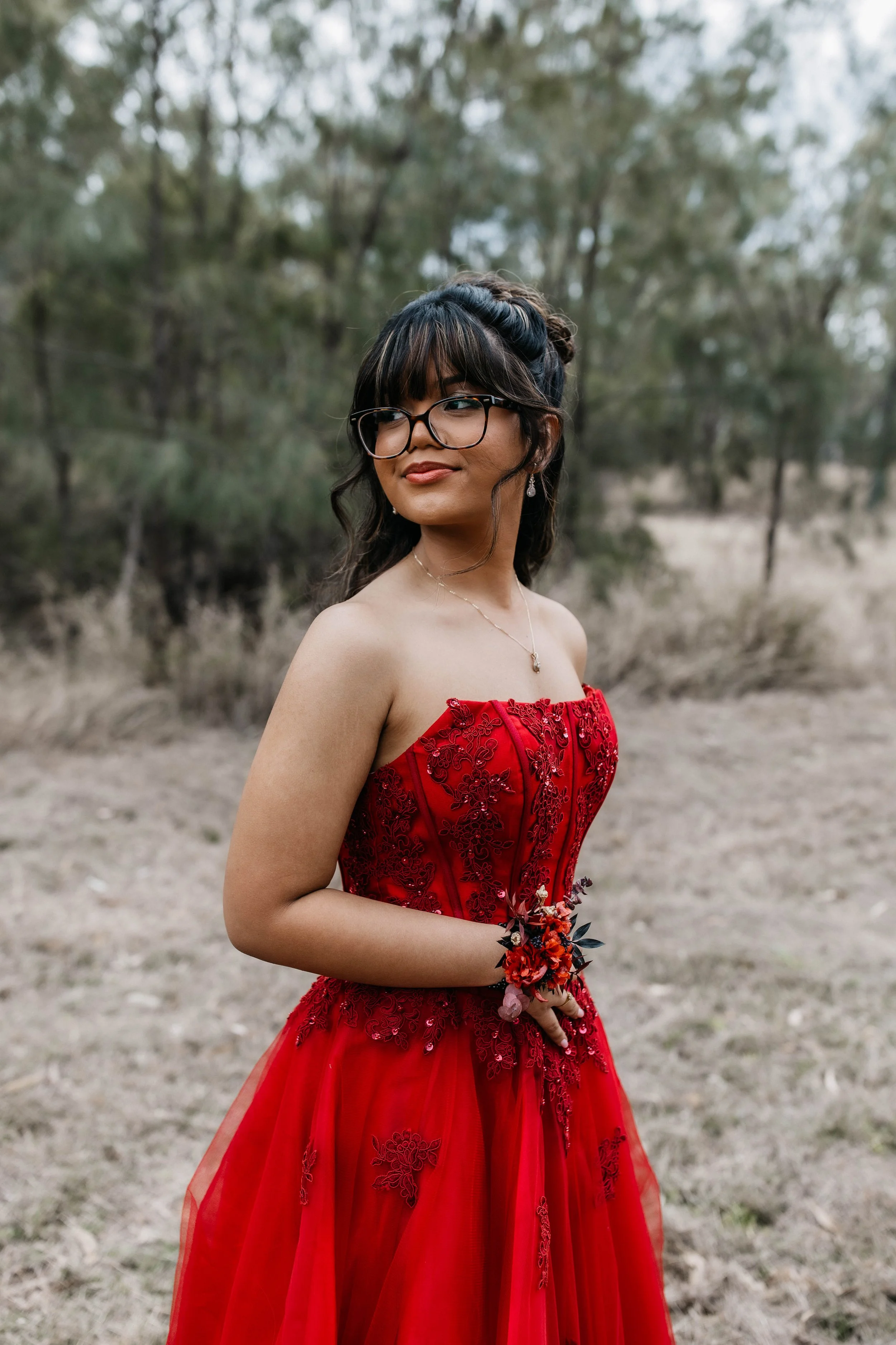 A woman in a red strapless dress with intricate embroidery poses outdoors with a blurred background of trees and dry grass. She has dark hair styled with curls and waves, wears glasses, and has earrings and a necklace. She holds a small bouquet of flowers in her hand.