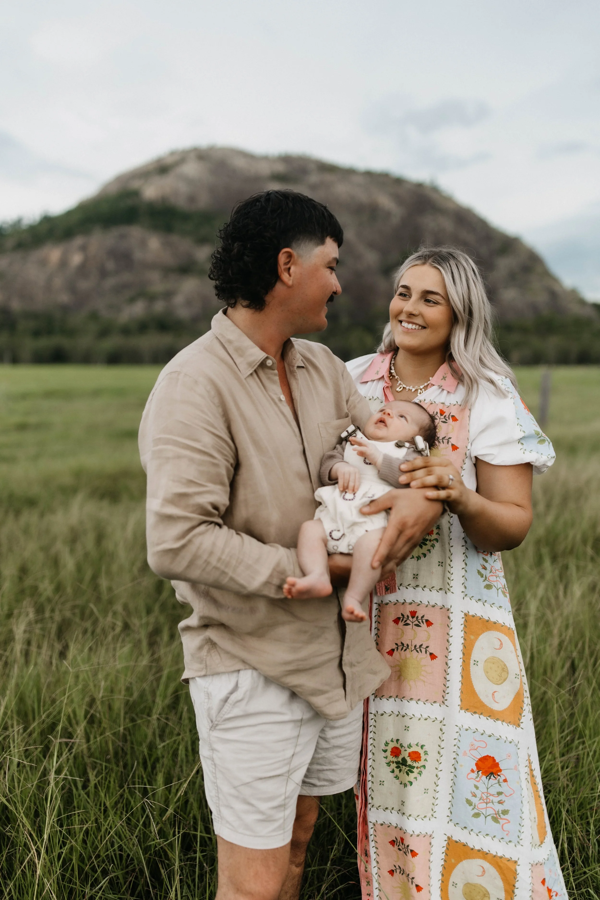 A man and woman standing in a grassy field with a mountain in the background. The man is holding a baby, and they are all smiling at each other.