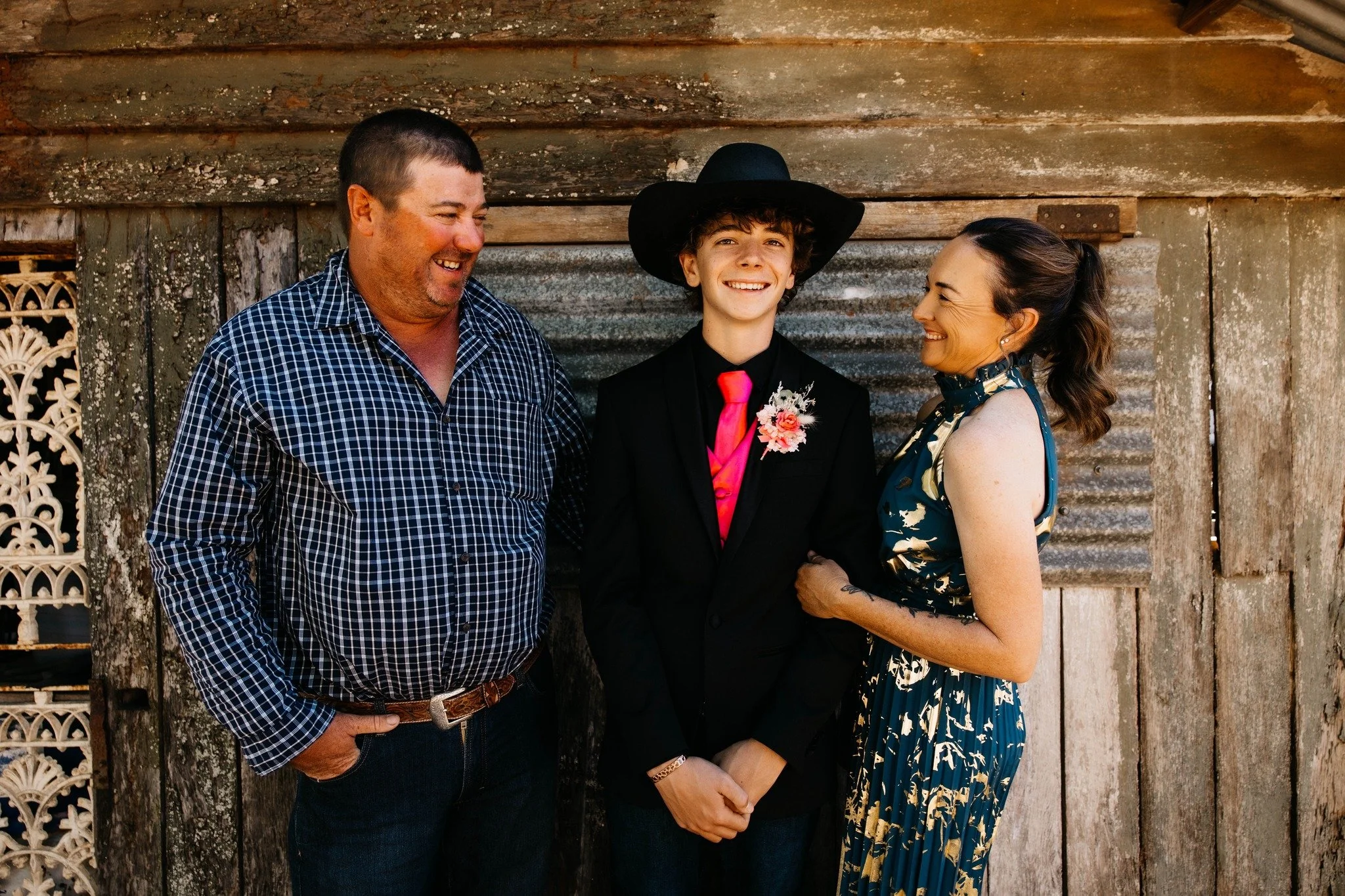 A group of three people, two adults and one teenager, smiling and looking at each other in front of an old rustic wooden wall. The teenager is wearing a black suit, pink tie, and a large black hat. One adult man is wearing a blue checkered shirt, and the woman is wearing a patterned dress with a scarf around her neck.
