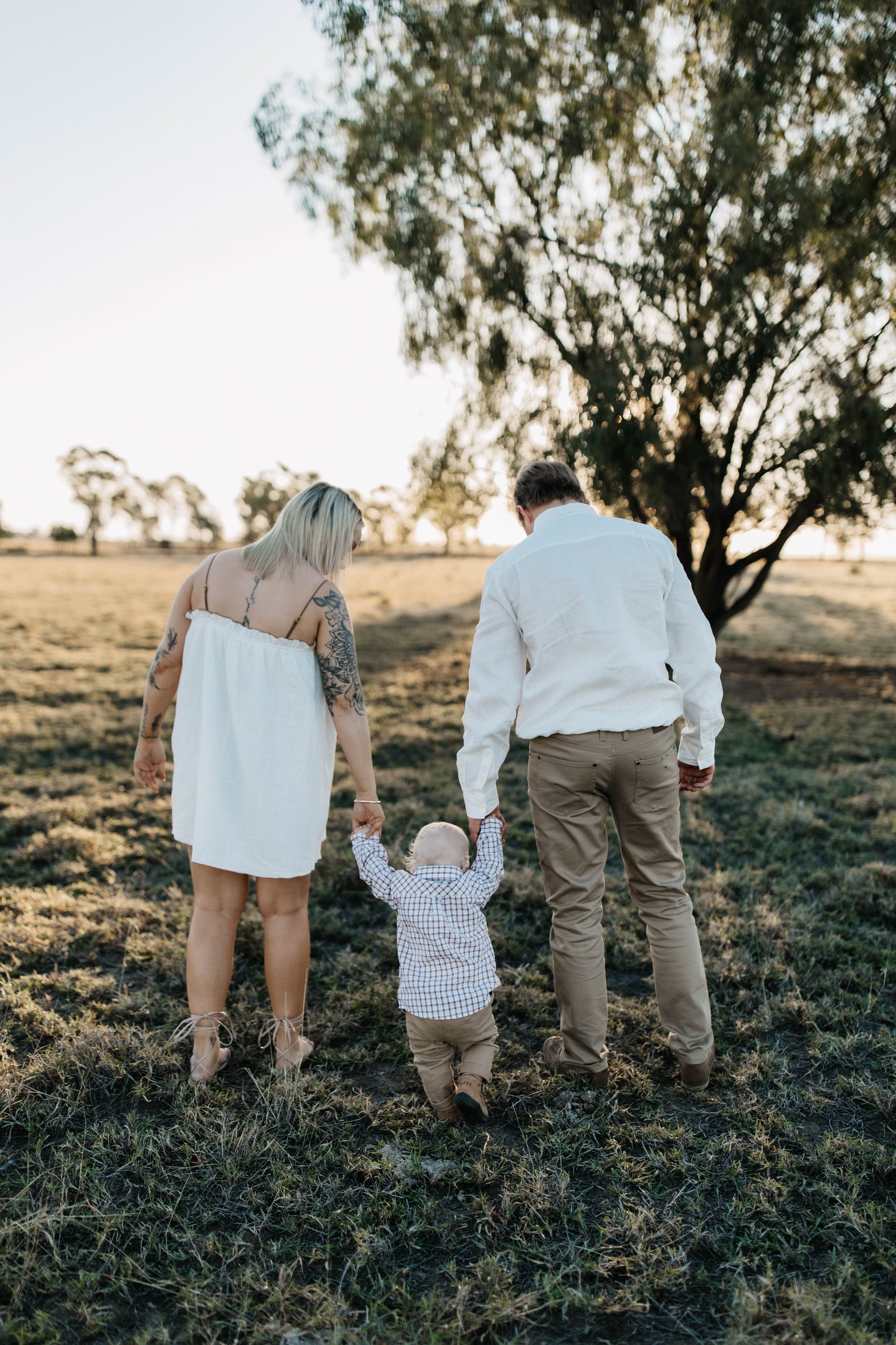 A family of three walking hand in hand through a grassy field during sunset, with trees in the background.