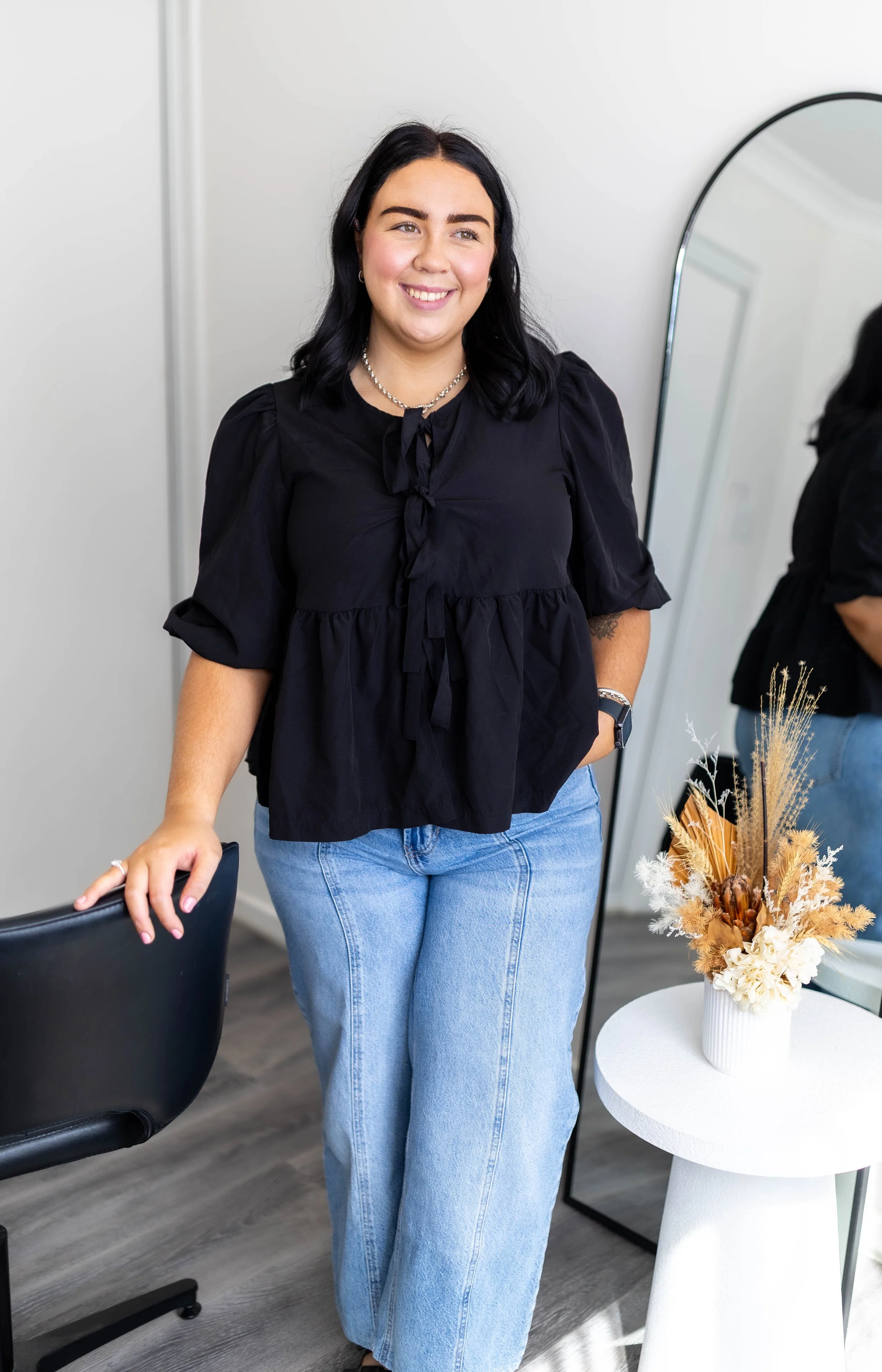 A woman with black hair smiling, wearing a black blouse and blue jeans, standing indoors near a round table with a floral arrangement, a mirror, and a black chair.
