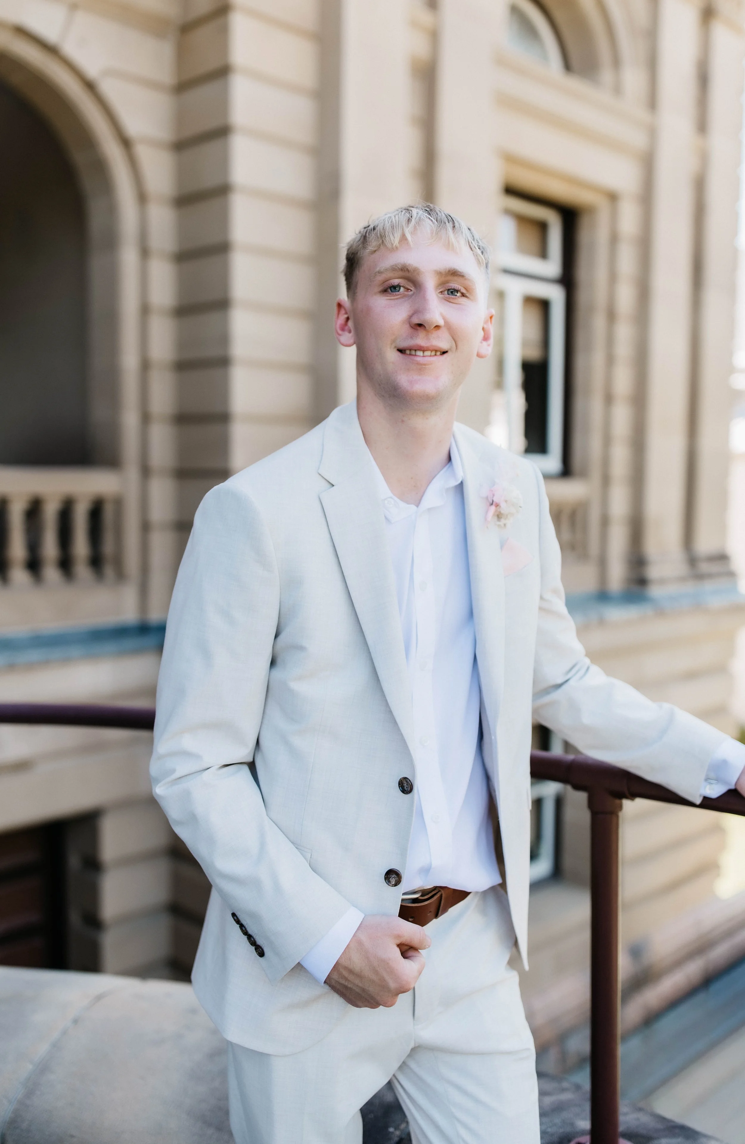 A young man in a white suit and white shirt standing on a balcony outside a building with beige stone walls and windows.