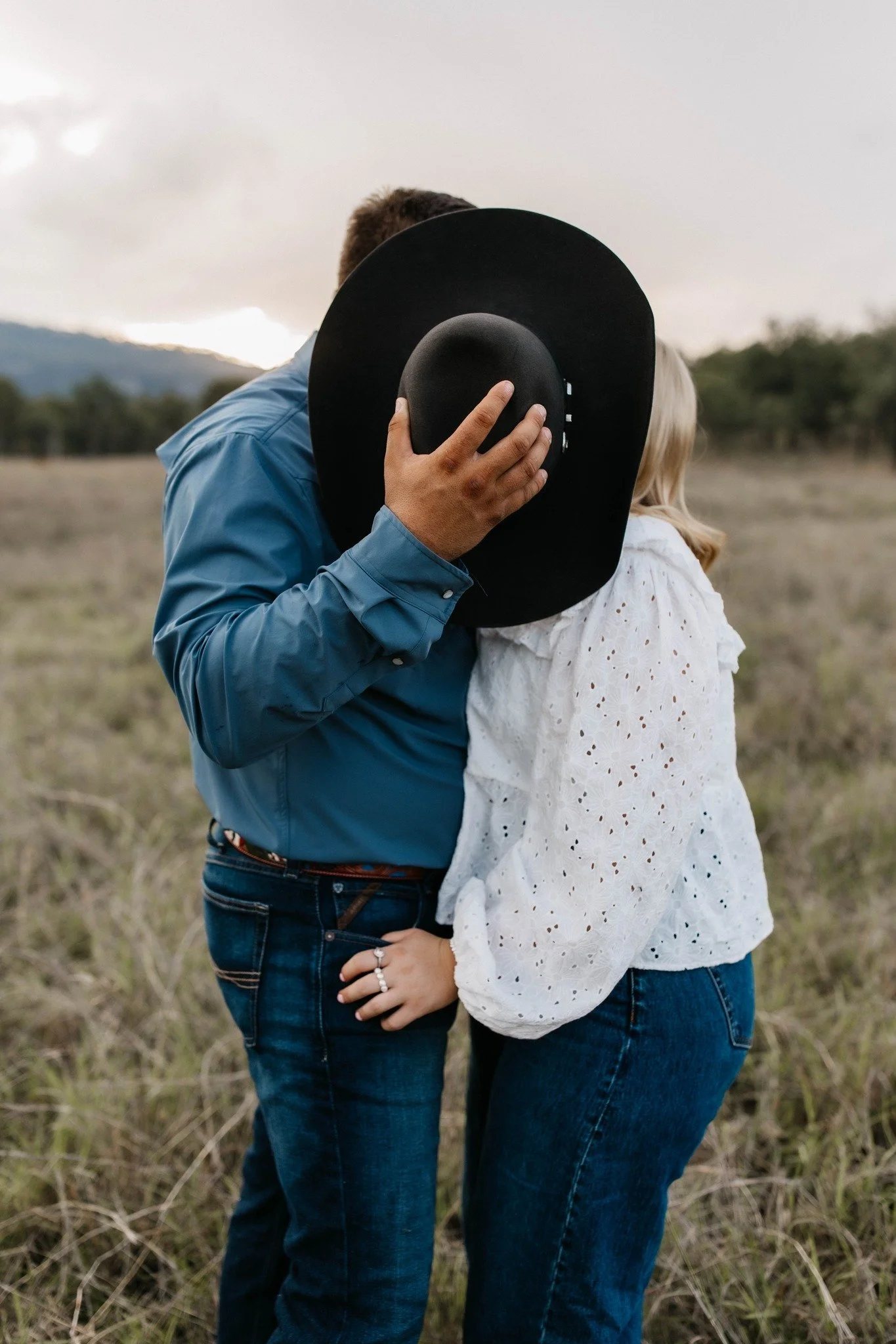 A man and woman sharing a kiss in a field, with the man holding a black hat and wearing a blue shirt, while the woman has blonde hair, a white blouse with eyelet details, and blue jeans.