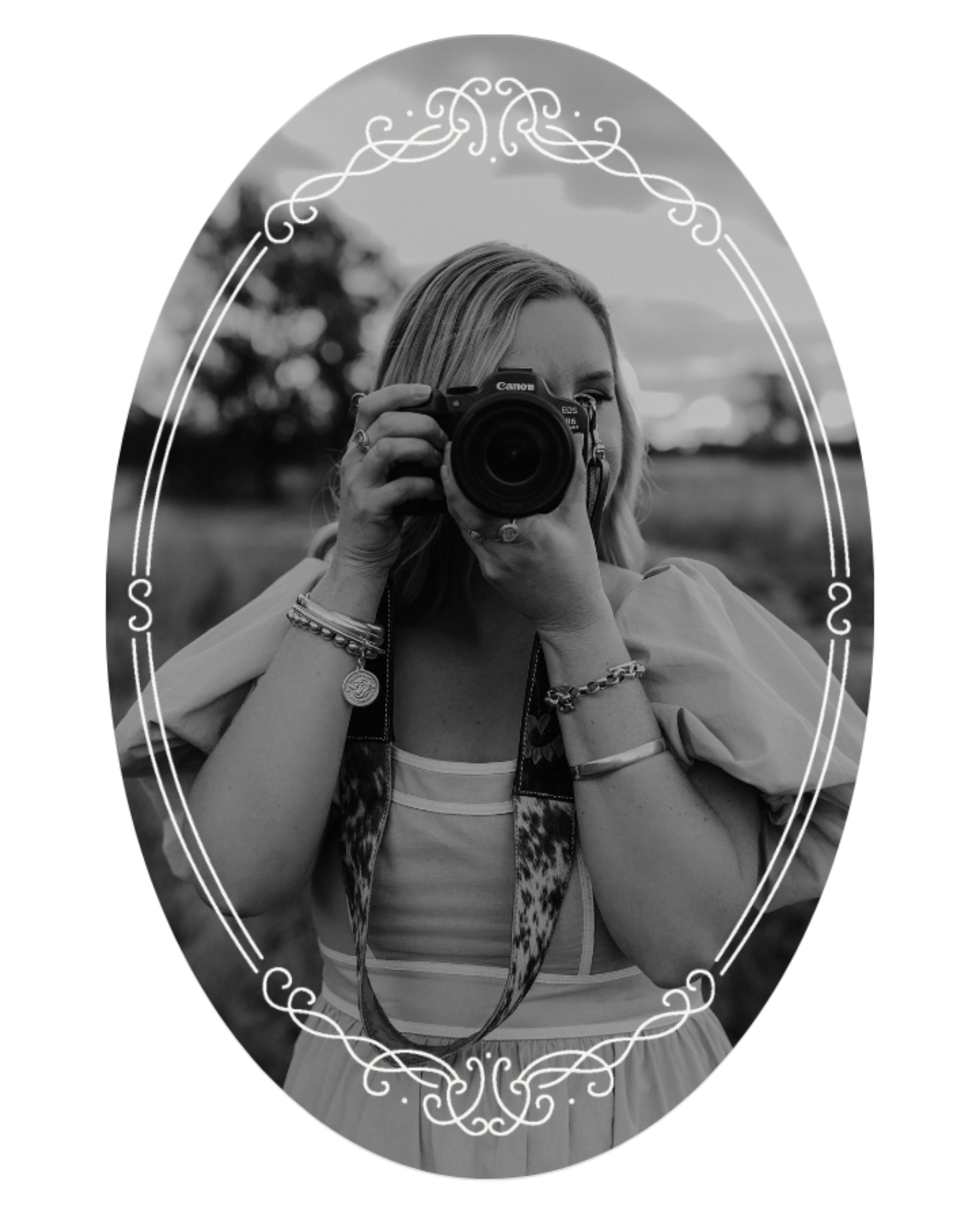 A woman with light hair holding a Canon camera up to her face, taking a self-portrait outdoors during daytime. She is wearing jewelry including bracelets, rings, and a necklace, and has a patterned scarf around her neck. The background is blurred with trees and sky visible.