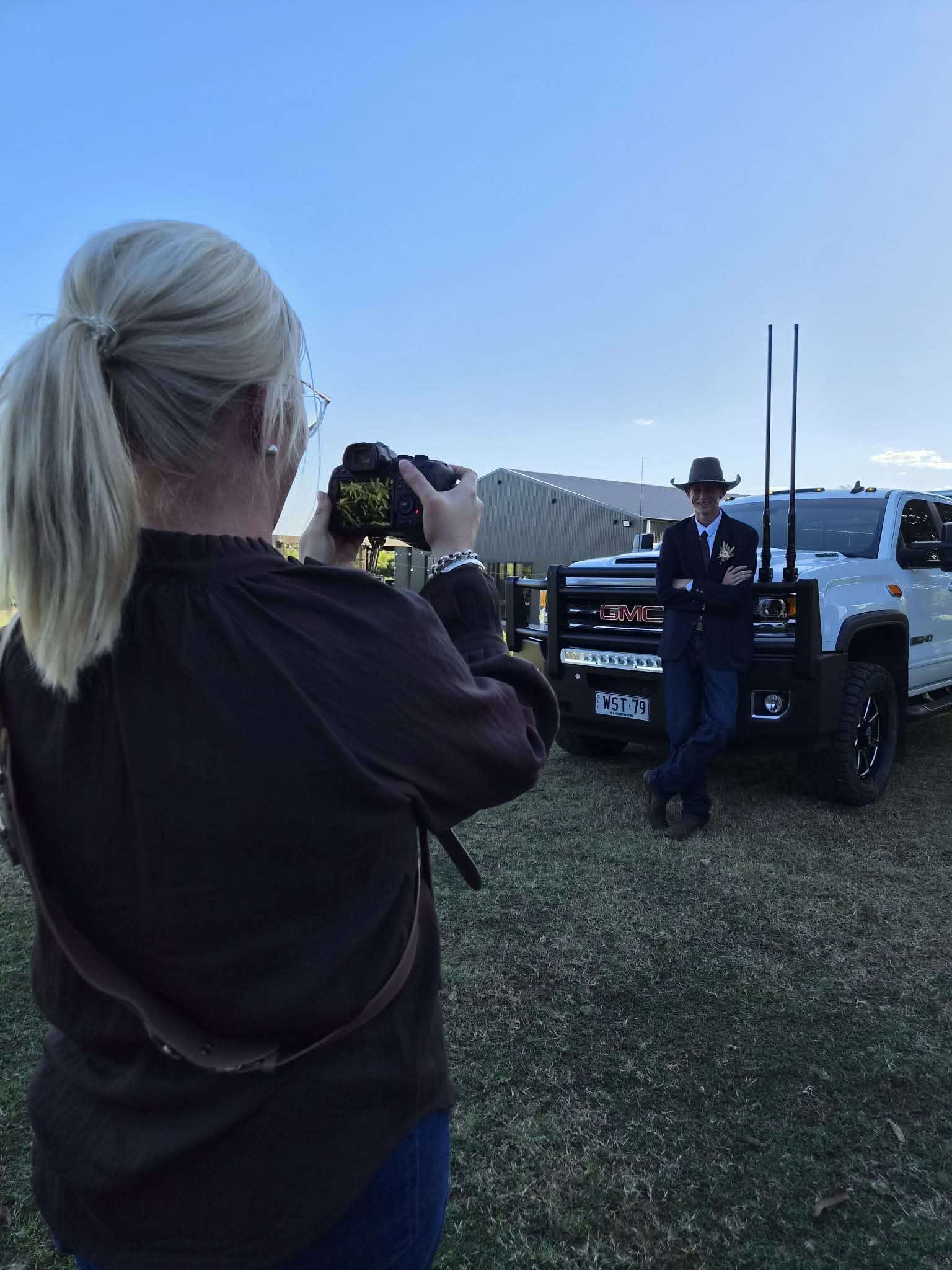 A woman with blonde hair tied back, wearing glasses and a black jacket, takes a photo of a man in a black suit, tie, and cowboy hat, standing in front of a GMC truck with two long antennas. They are outdoors on grass, with a large metal building in the background and the sky is clear and blue.
