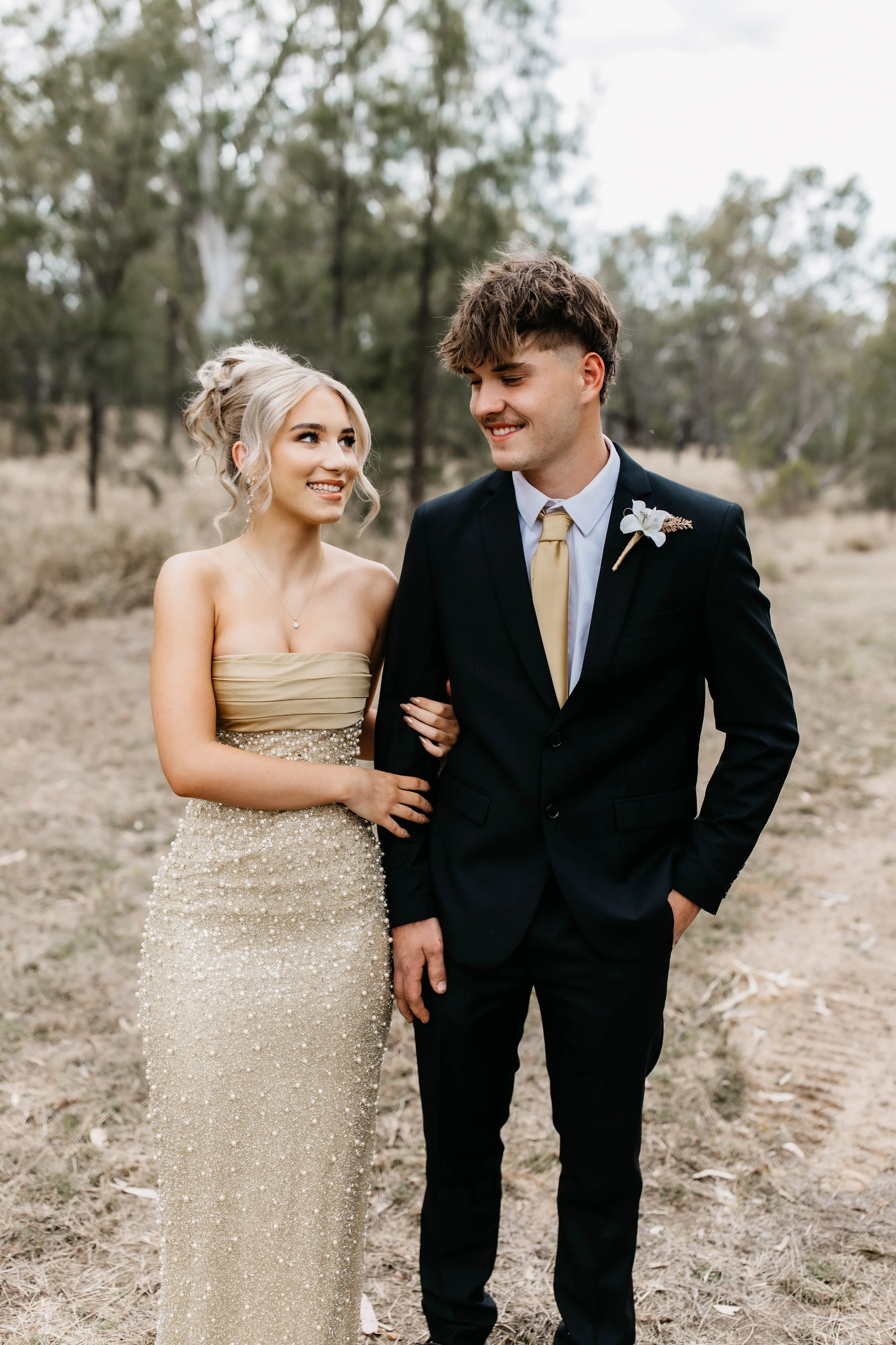 A bride and groom standing outdoors in a natural setting with trees, smiling at each other; the bride in a strapless, sparkling, gold-colored dress, and the groom in a dark suit with a boutonnière.