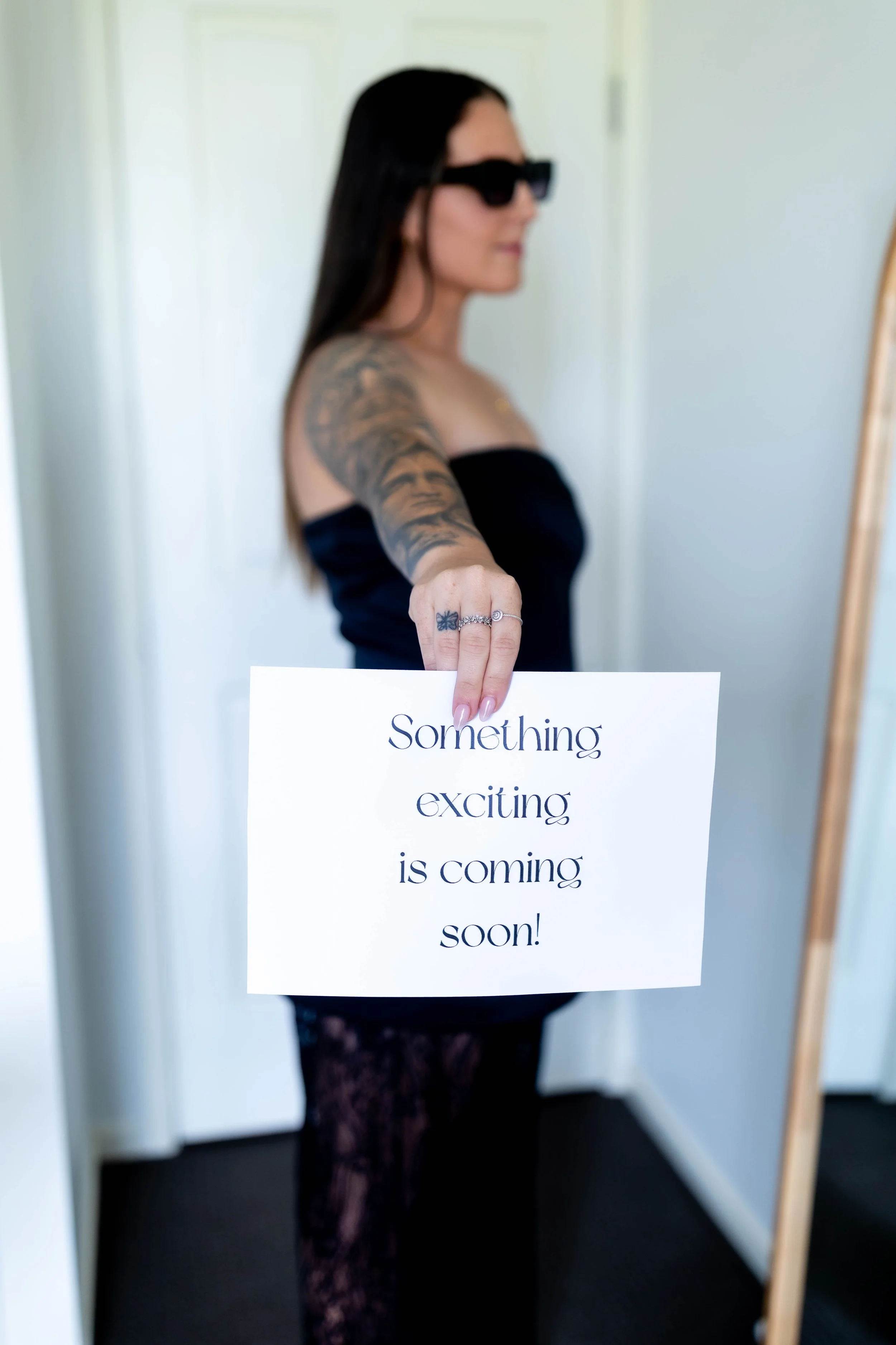 A woman with dark hair, sunglasses, tattoos, wearing a strapless black top, holding a sign that reads 'Something exciting is coming soon!' in an indoor setting.