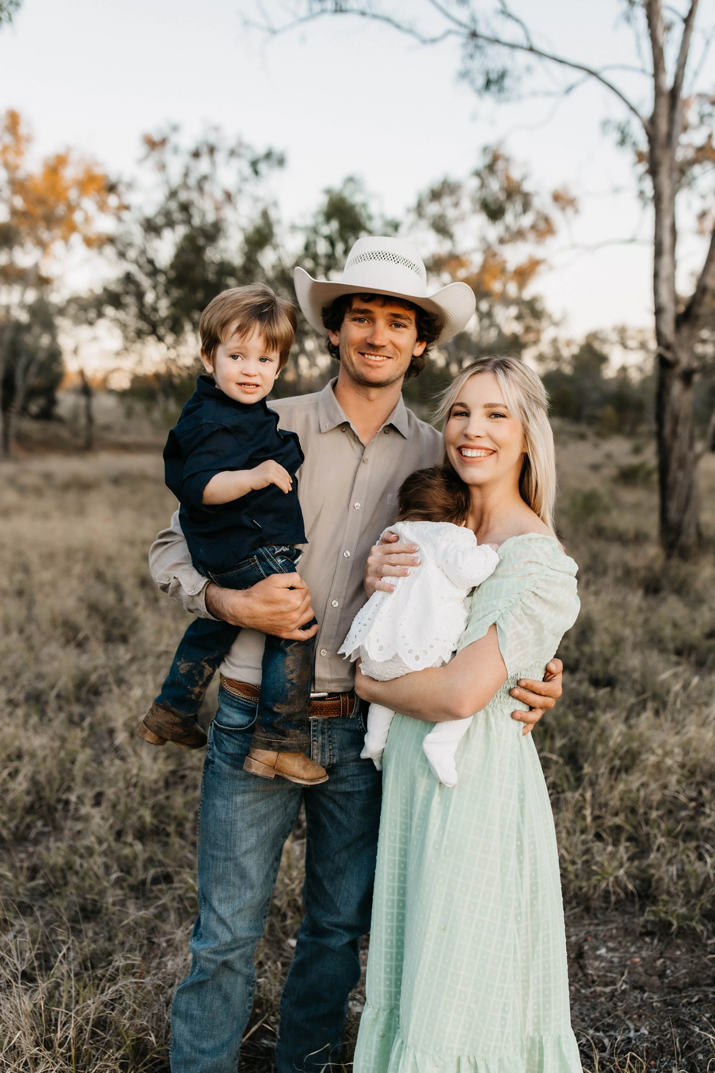 A family of four standing outdoors in a field with trees in the background, smiling and holding their children, one boy and one girl, during sunset.