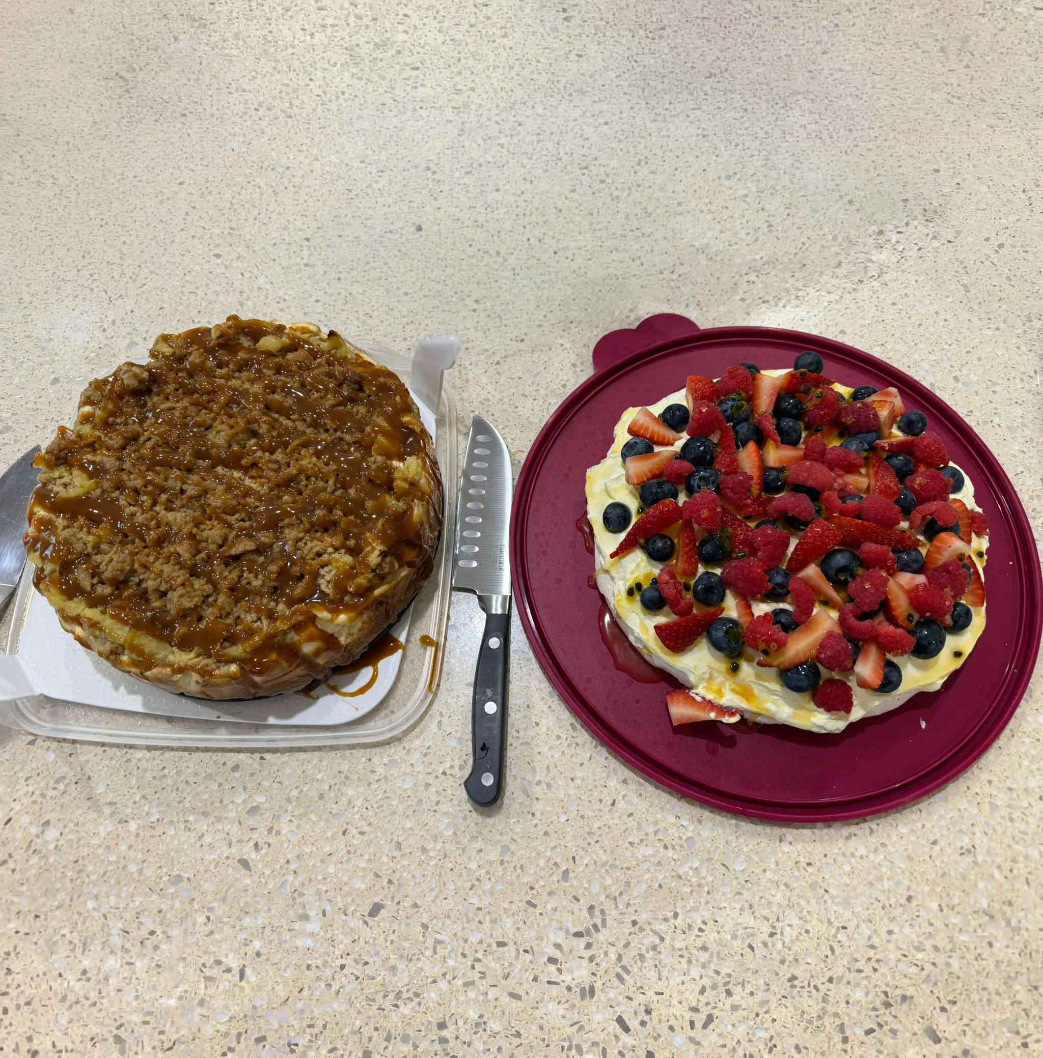 Two types of desserts on a speckled beige countertop: a caramel apple crumble pie on the left and a fruit-topped cheesecake on a red plate on the right.