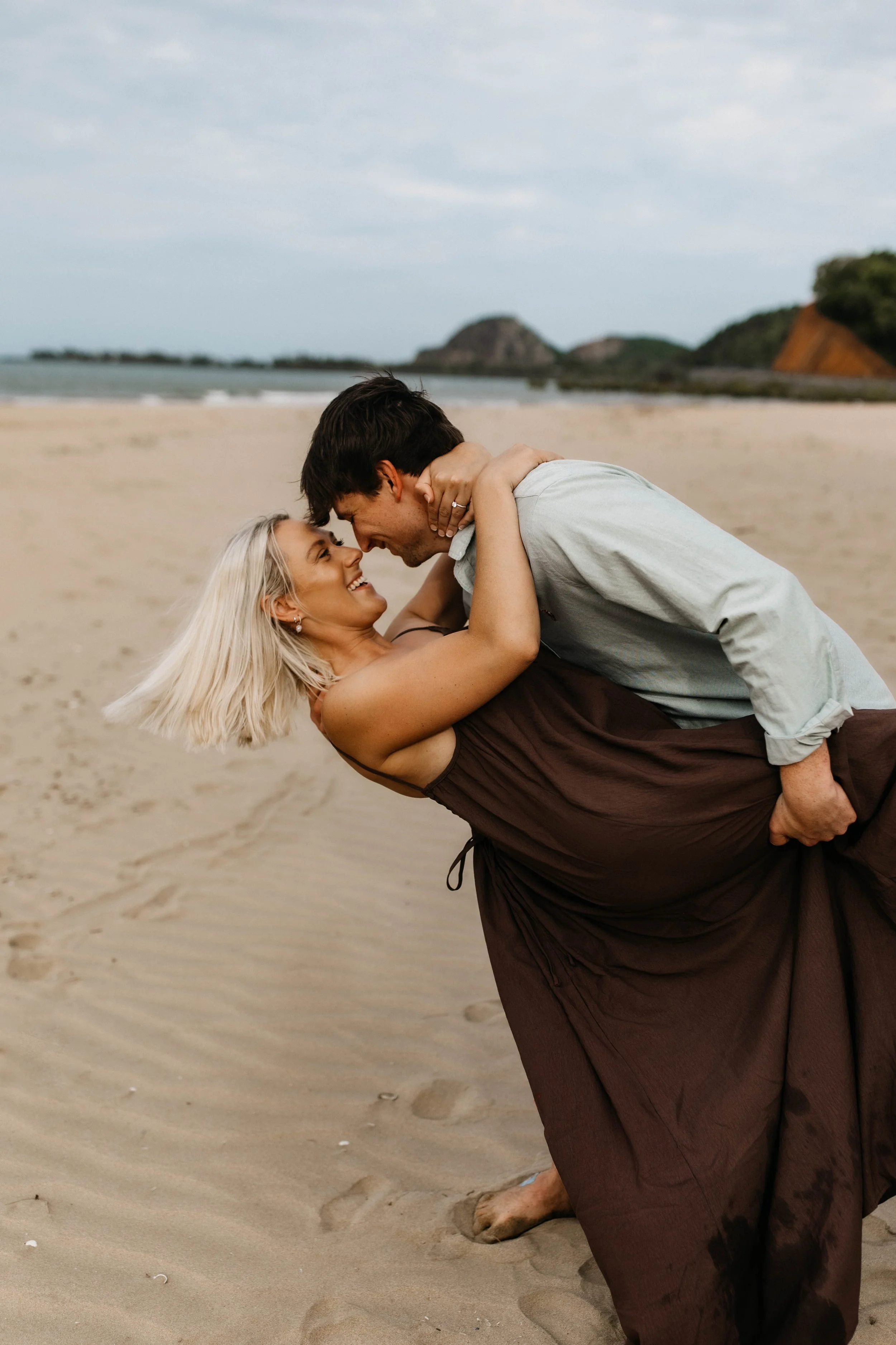 A couple on the beach, with the man dipping the woman who is smiling, in a loving embrace. The woman has blonde hair and the man has dark hair, and they are both dressed casually. The background shows sandy shore, ocean, and distant rocky islands under a cloudy sky.
