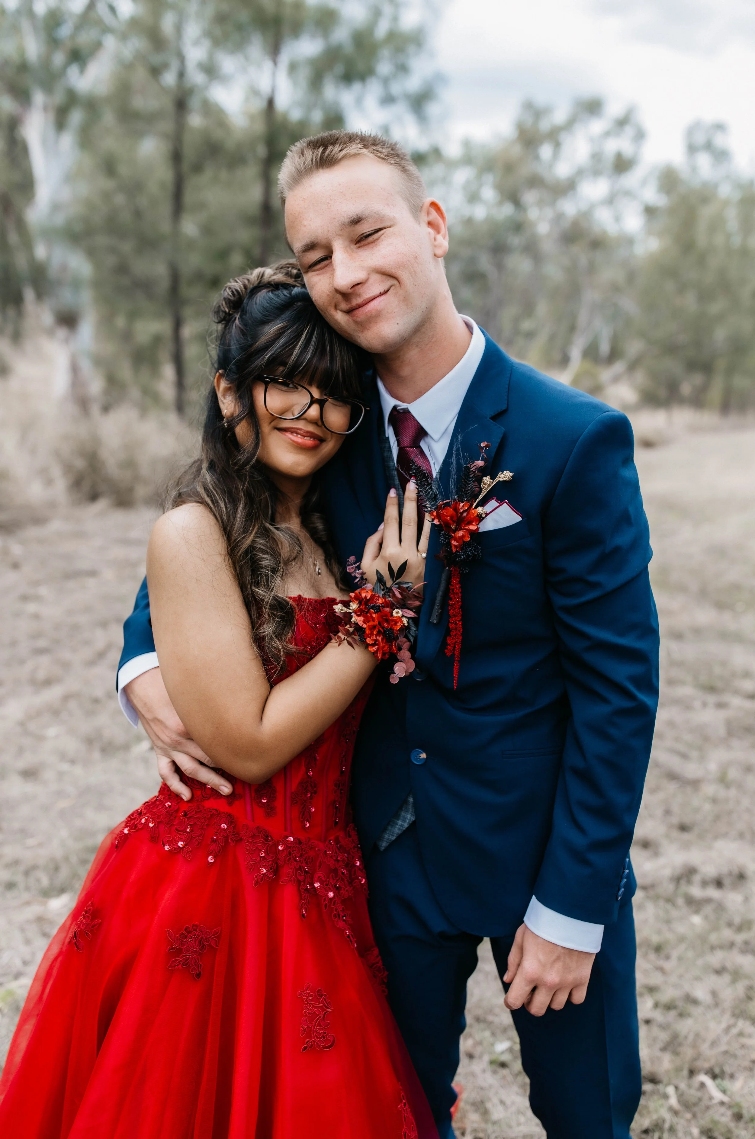 A young man and woman embracing outdoors, the woman wearing a red dress with floral embroidery and glasses, the man in a blue suit with a boutonniere, both smiling with eyes closed.