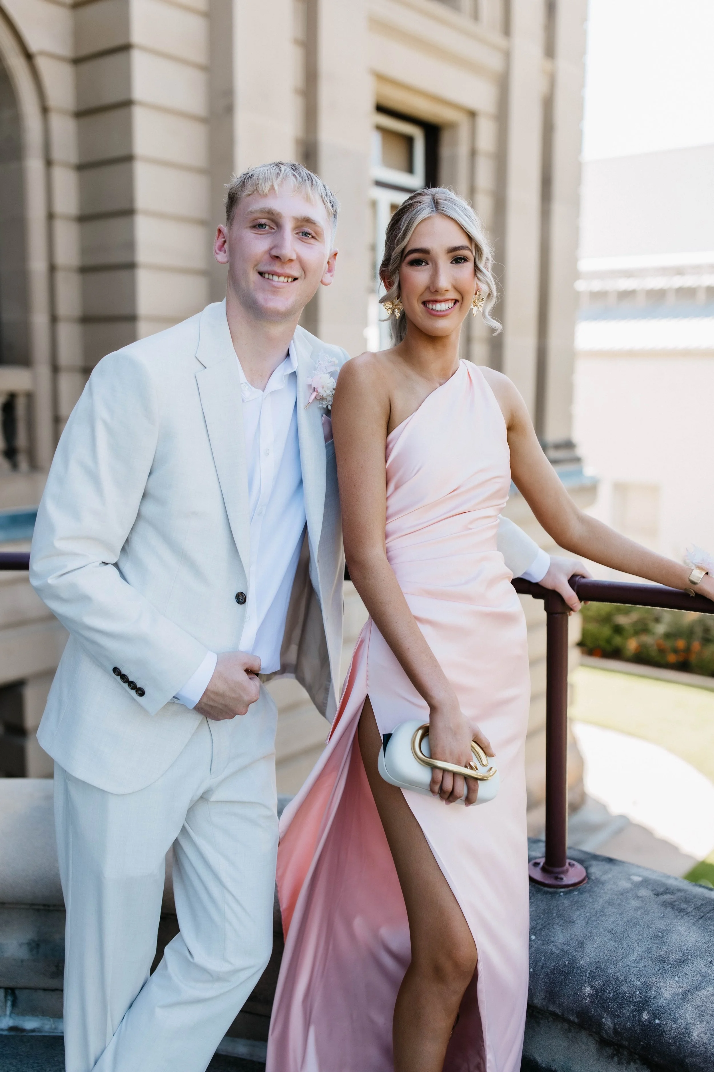 A smiling young man in a white suit and a young woman in a pink dress standing outdoors by a stone staircase, with a building in the background, during daytime.