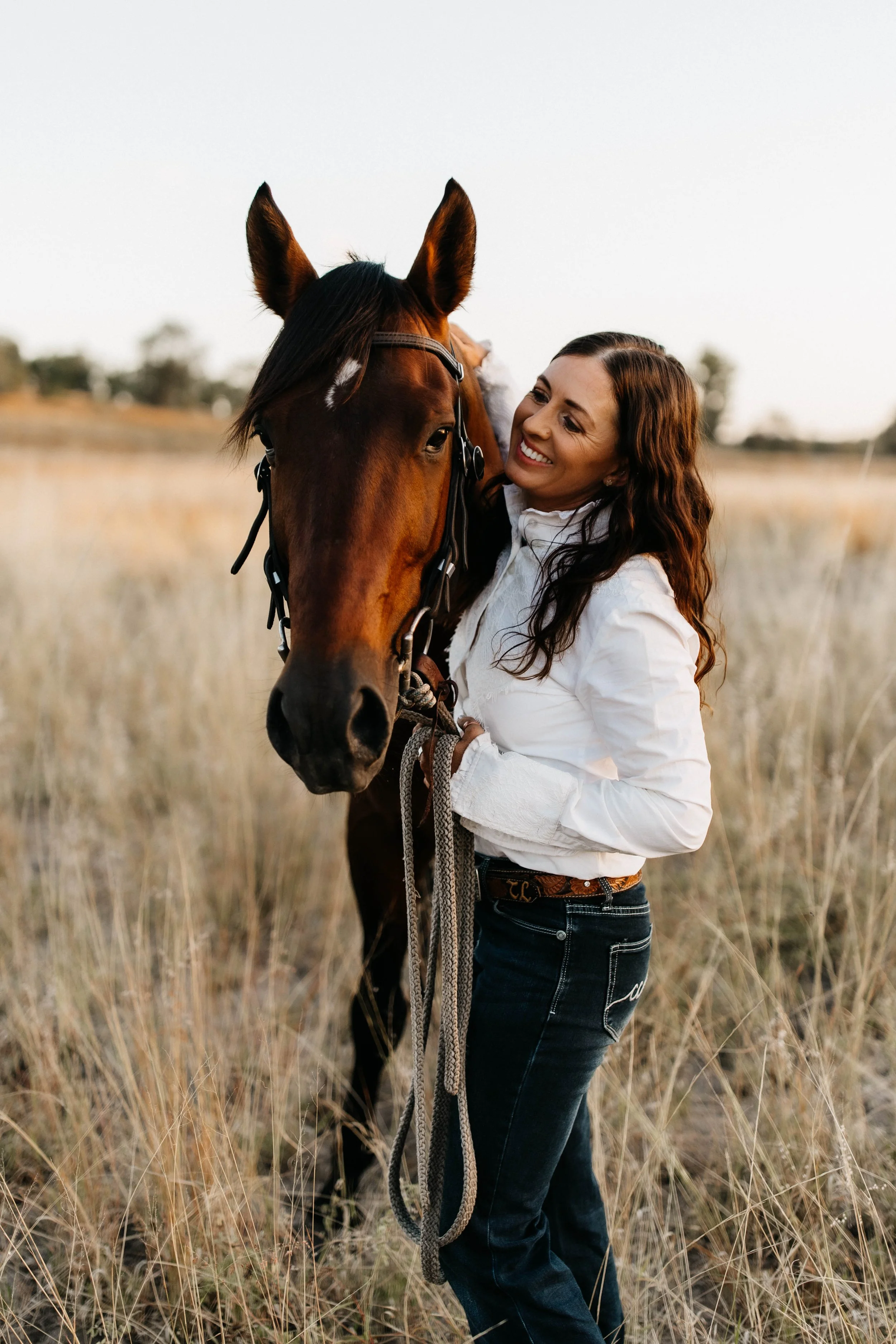 A woman with long brown hair smiling and hugging a brown horse in a field of tall, dry grass during sunset.