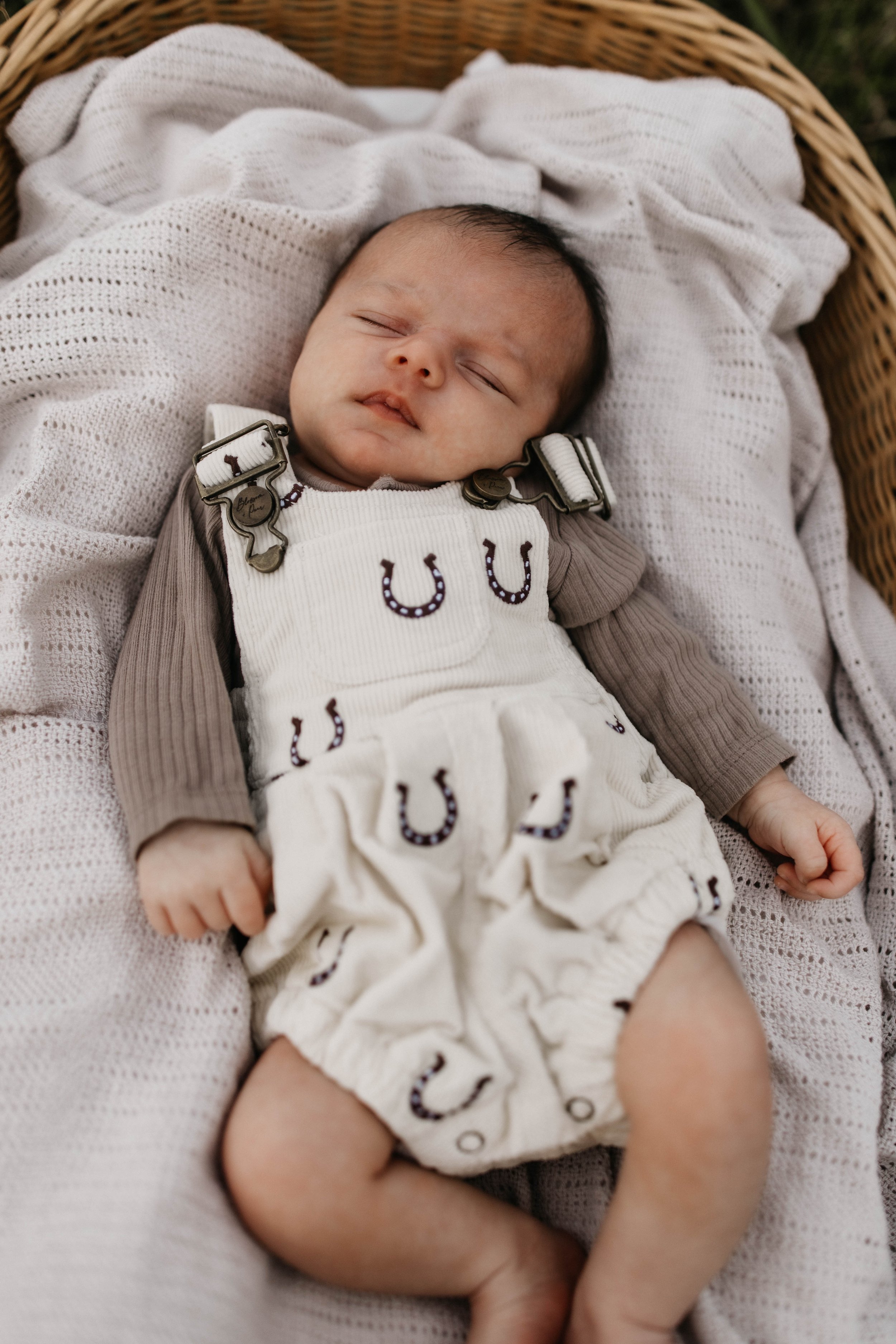 A sleeping baby lying in a wicker basket on a white, knitted blanket, with a person holding their leg.