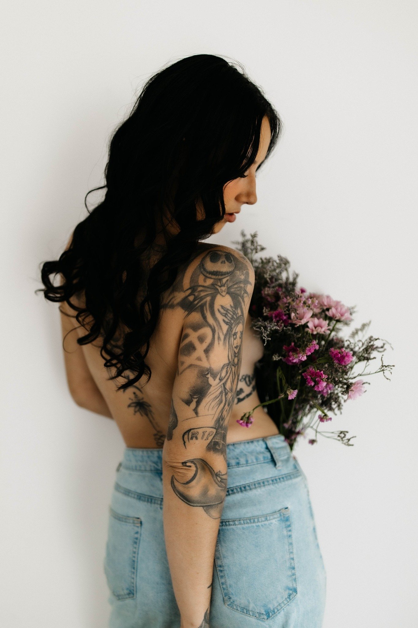 A young woman with long black curly hair, tattoos on her left arm and shoulder, holding a bouquet of pink and purple flowers, wearing light blue jeans, standing against a plain white wall.