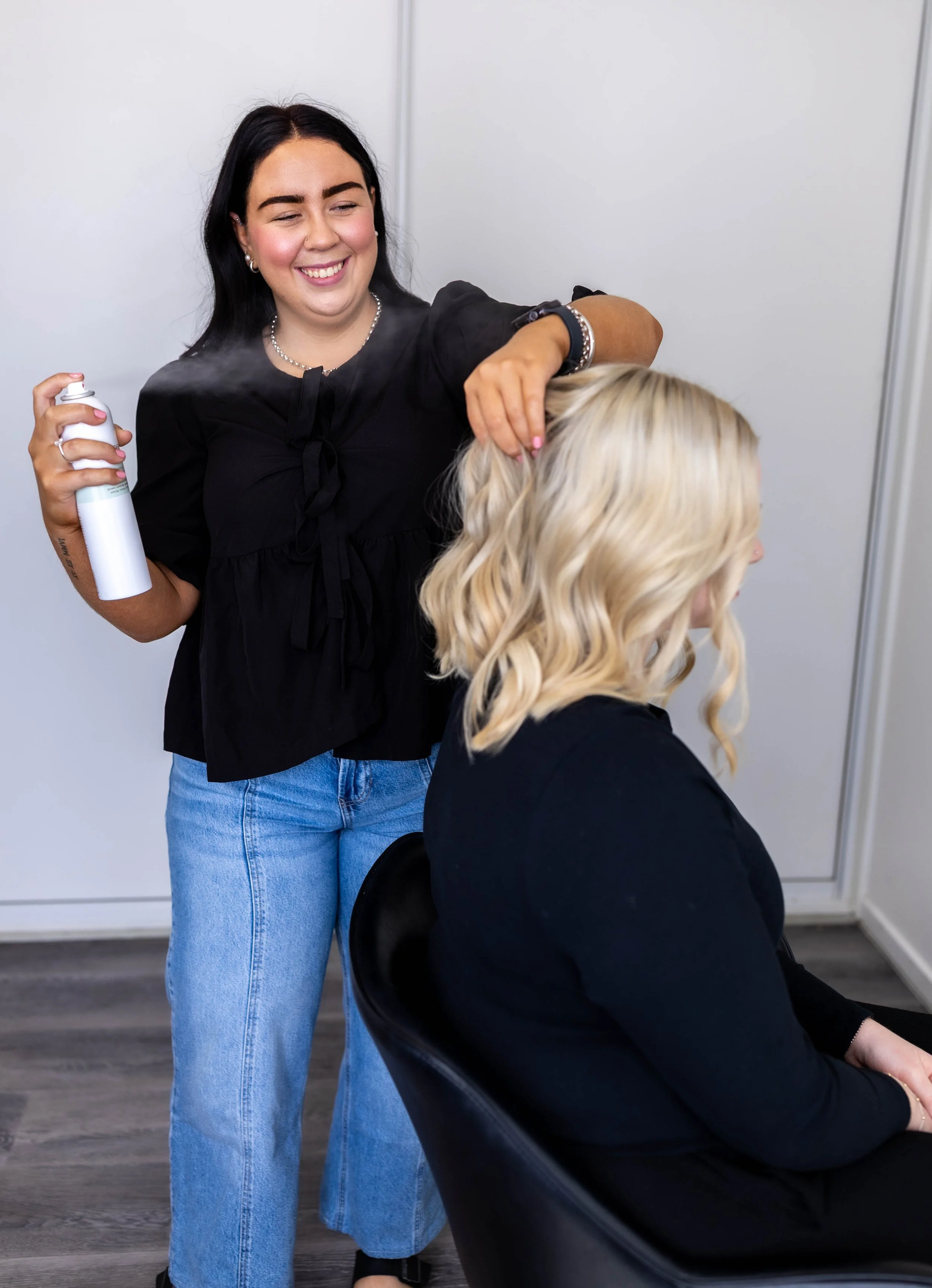 Hair stylist spray hair with aerosol can while smiling at client seated in salon chair.
