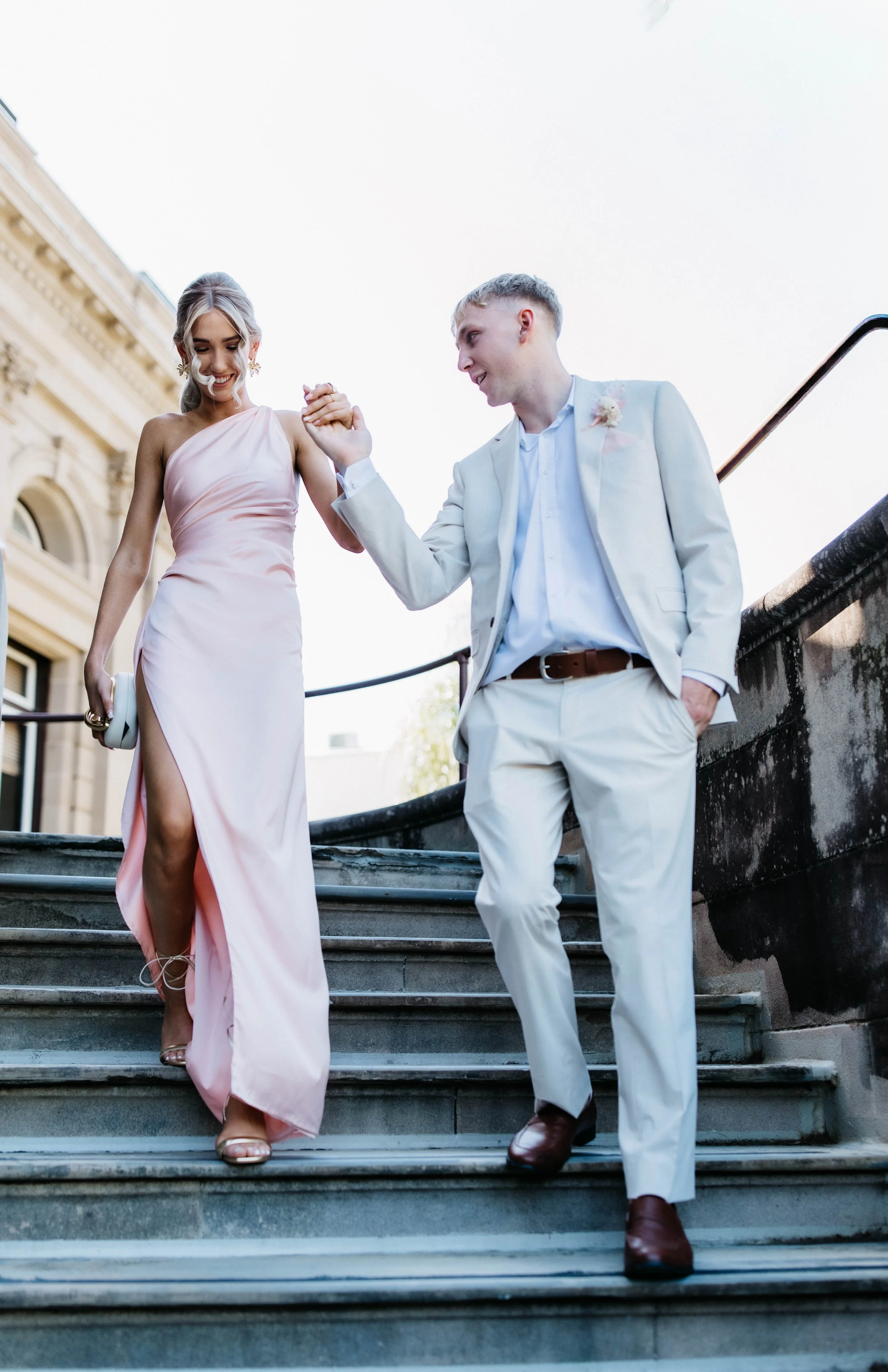 A loving couple is walking down the stairs outdoors on a sunny day. The woman is wearing a pink satin evening gown with a thigh-high slit, and the man is dressed in a white suit with a boutonniere. They are holding hands and smiling at each other.