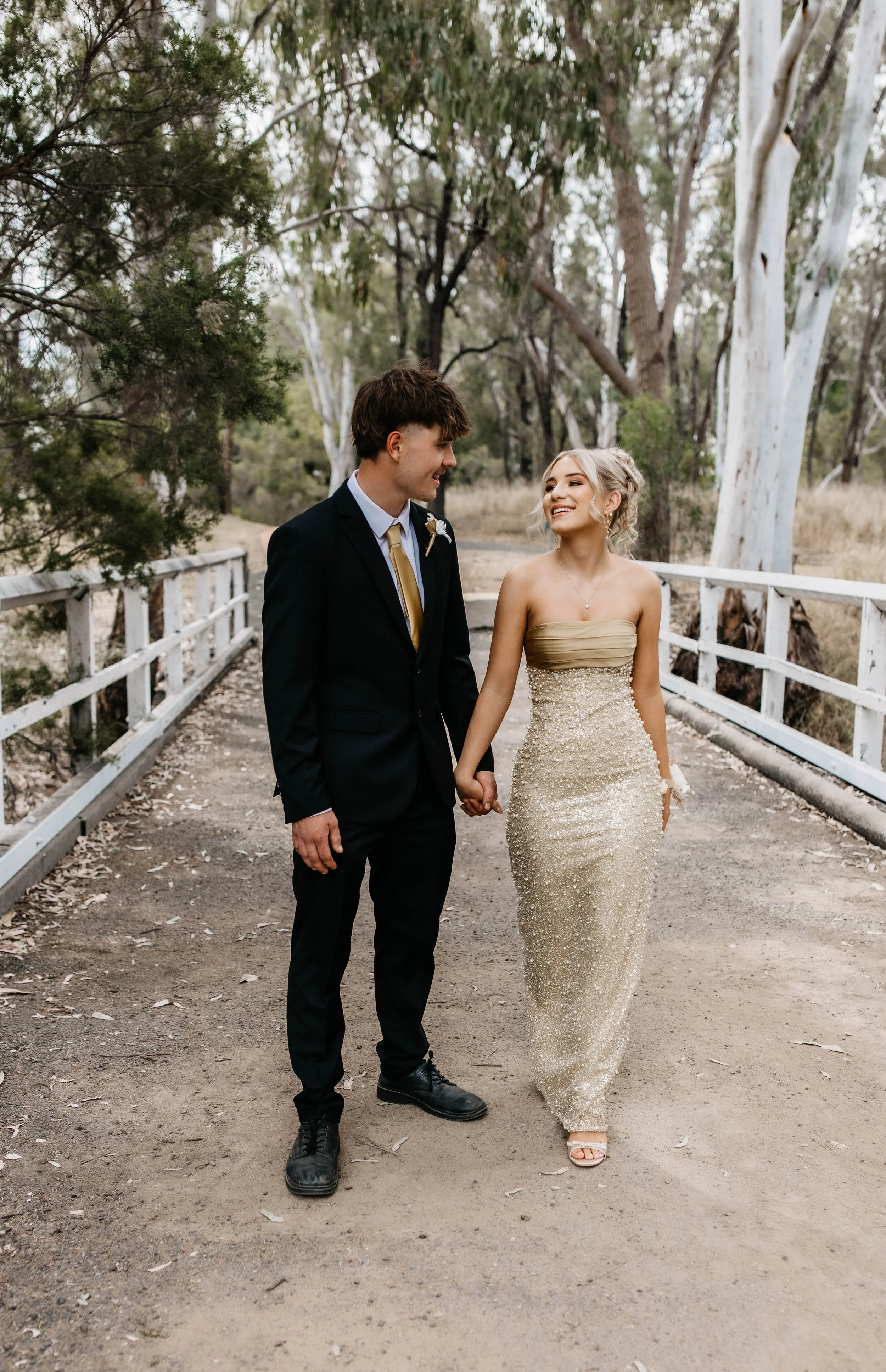 A young couple dressed in formal wedding attire holding hands and walking on a dirt path through a wooded area, smiling at each other.