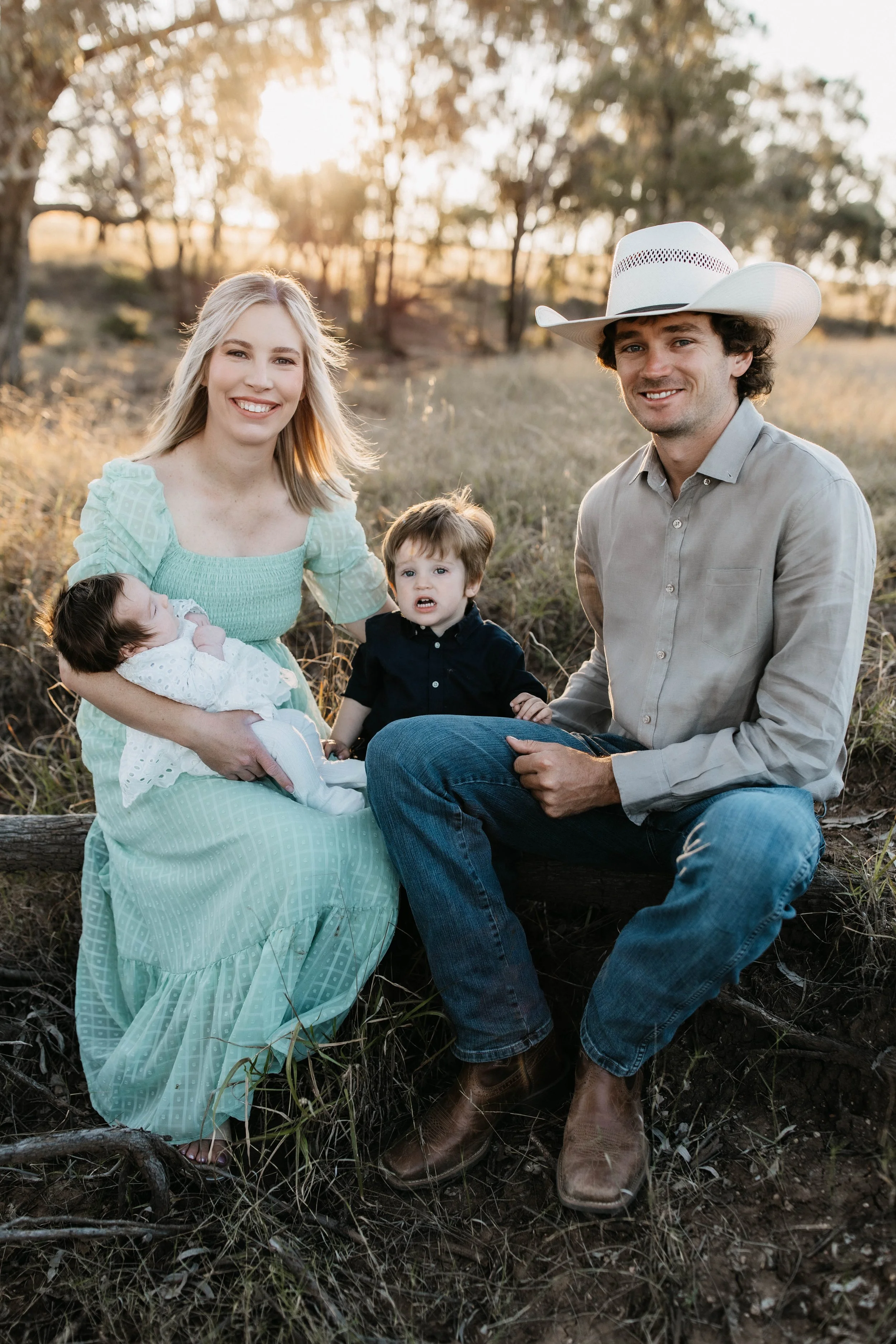 A smiling family of four outdoors in a rural setting during sunset. The mother holds a sleeping baby girl, the father wears a cowboy hat, and two young children, a girl and a boy, are sitting on a log.