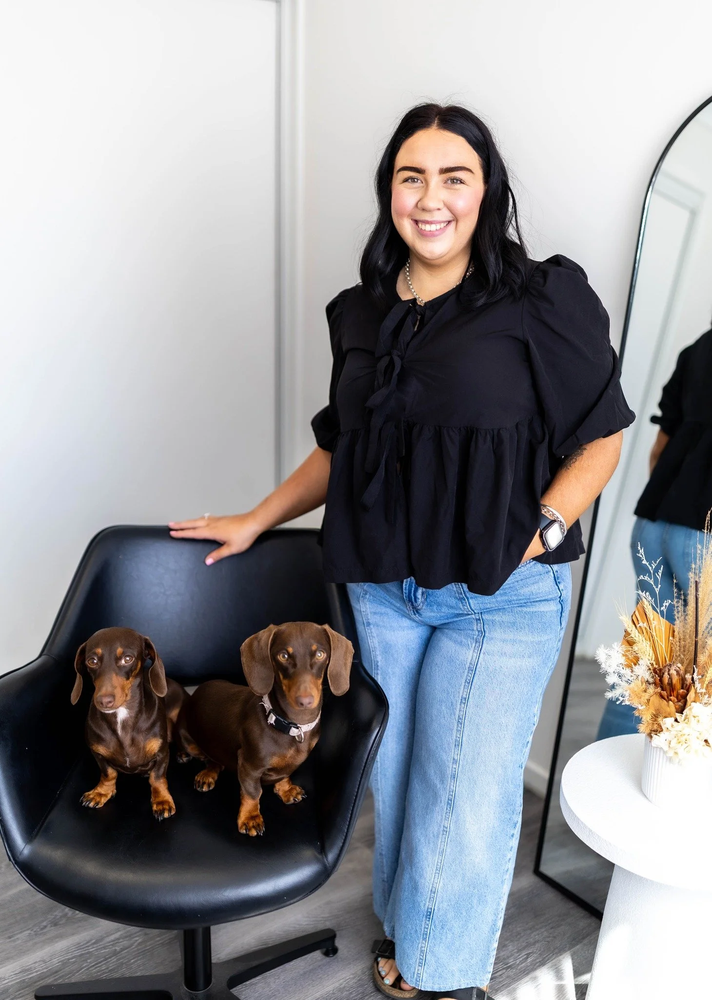 A woman with black hair, wearing a black blouse and blue jeans, standing indoors next to a black chair with two brown dachshunds sitting on it. There is a white side table with a vase of dried flowers nearby.