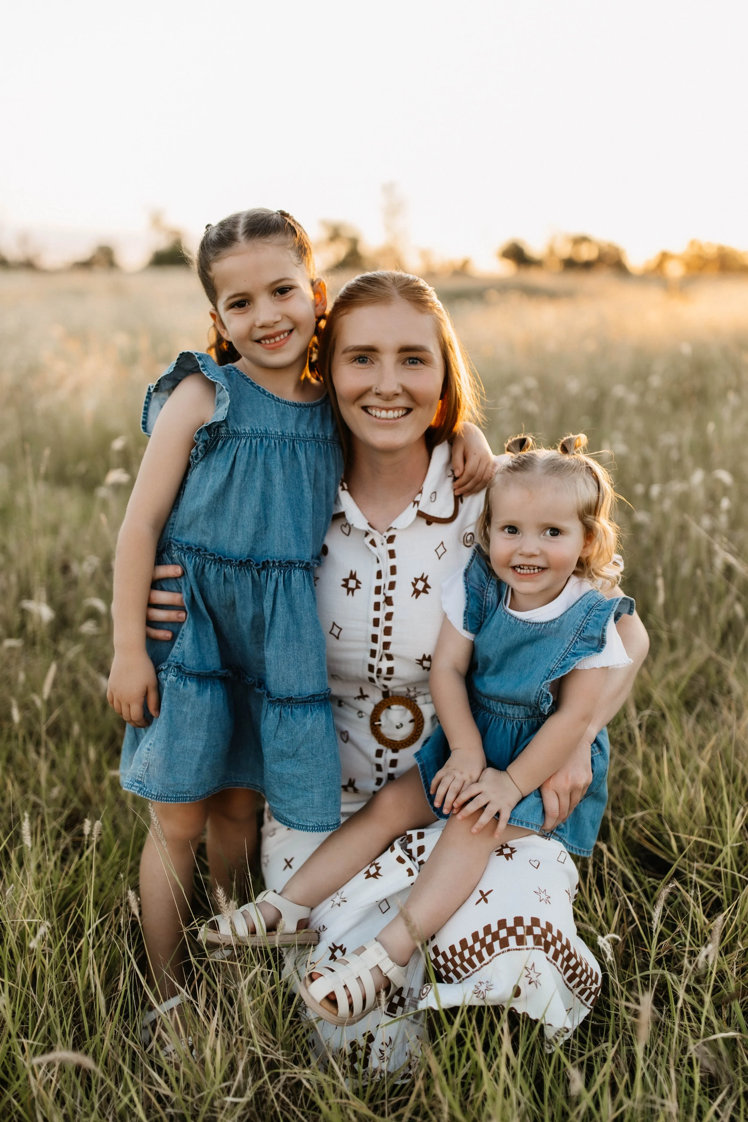 A woman and two young girls smiling in a grassy field at sunset. The woman is kneeling, and the girls are standing and sitting, all wearing casual summer clothes.