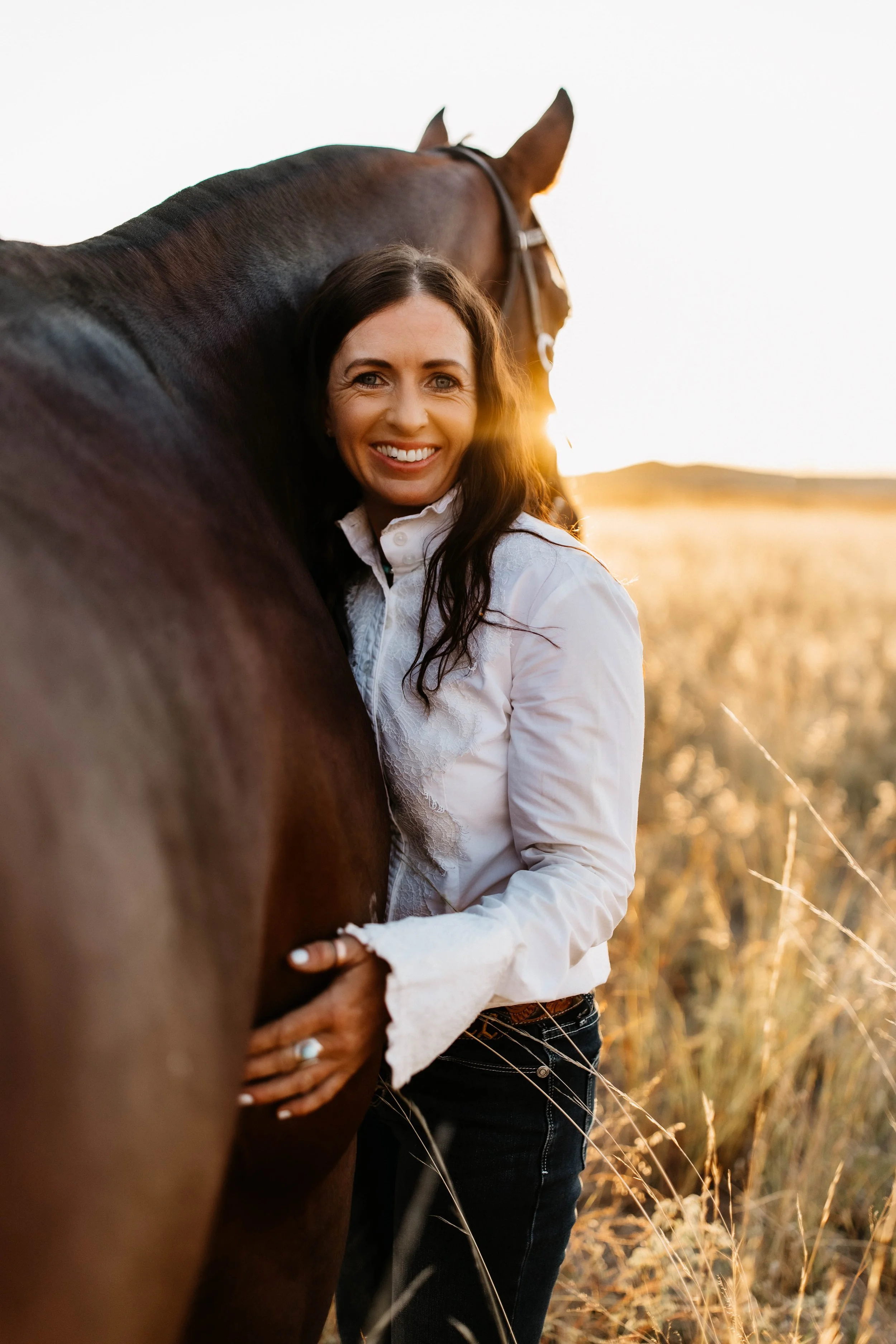A woman with long dark hair smiling and hugging a brown horse during sunset in an open field with tall grass.