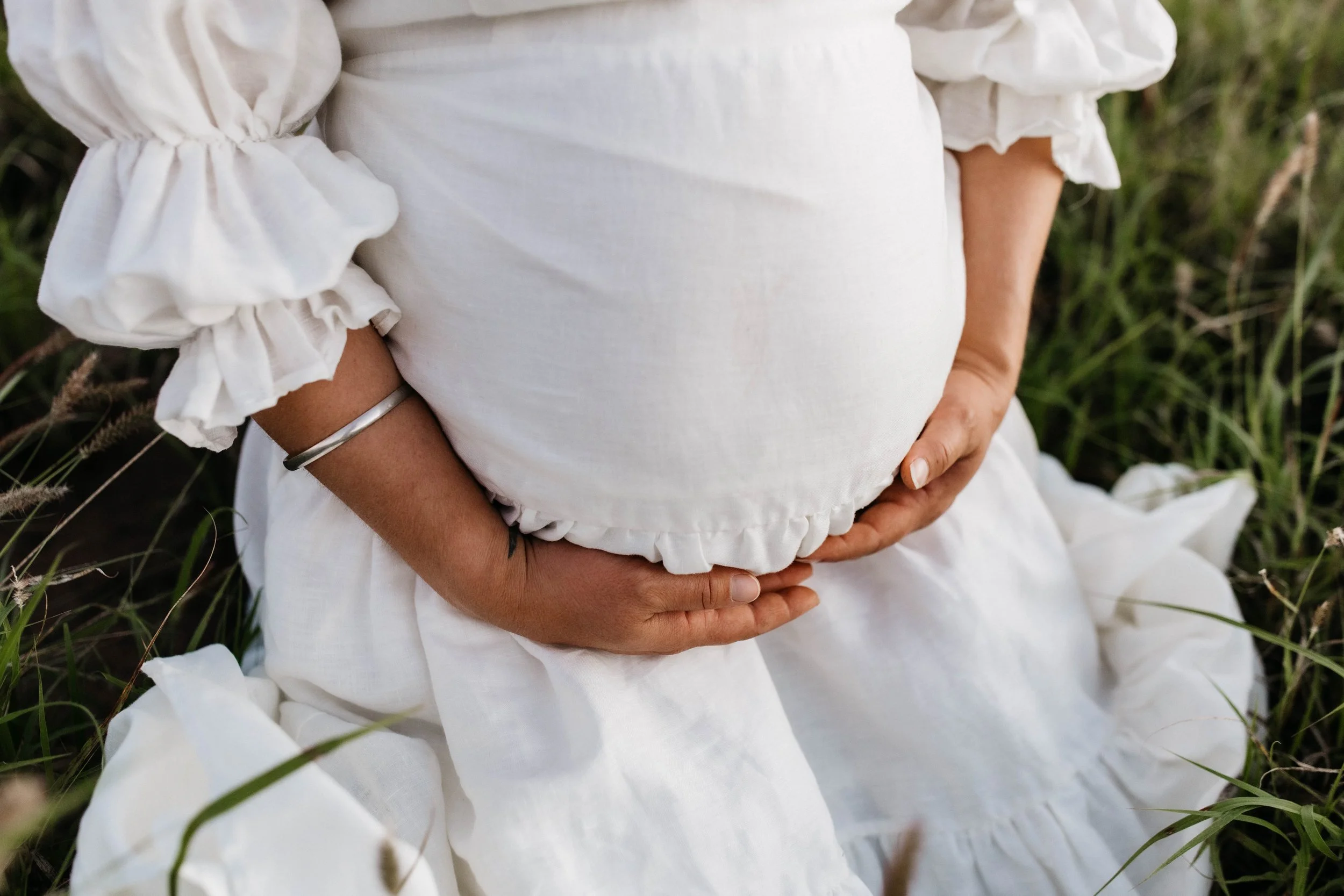 A pregnant woman wearing a white dress gently holding her belly while sitting outdoors among tall grass.