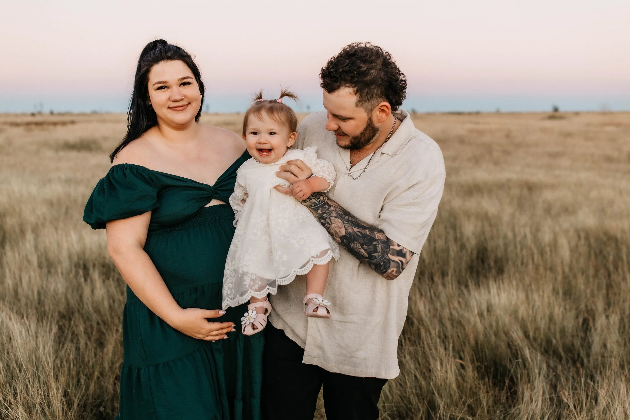 A family of three standing in a field at sunset, with a pregnant woman, a man holding a smiling baby girl, and a vast landscape in the background.