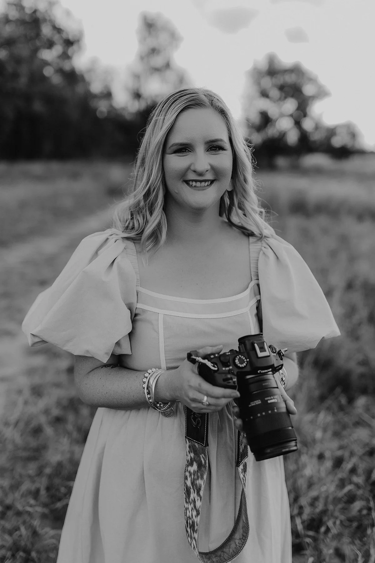 A smiling woman with blonde hair holding a professional camera outdoors in a grassy field with trees in the background.