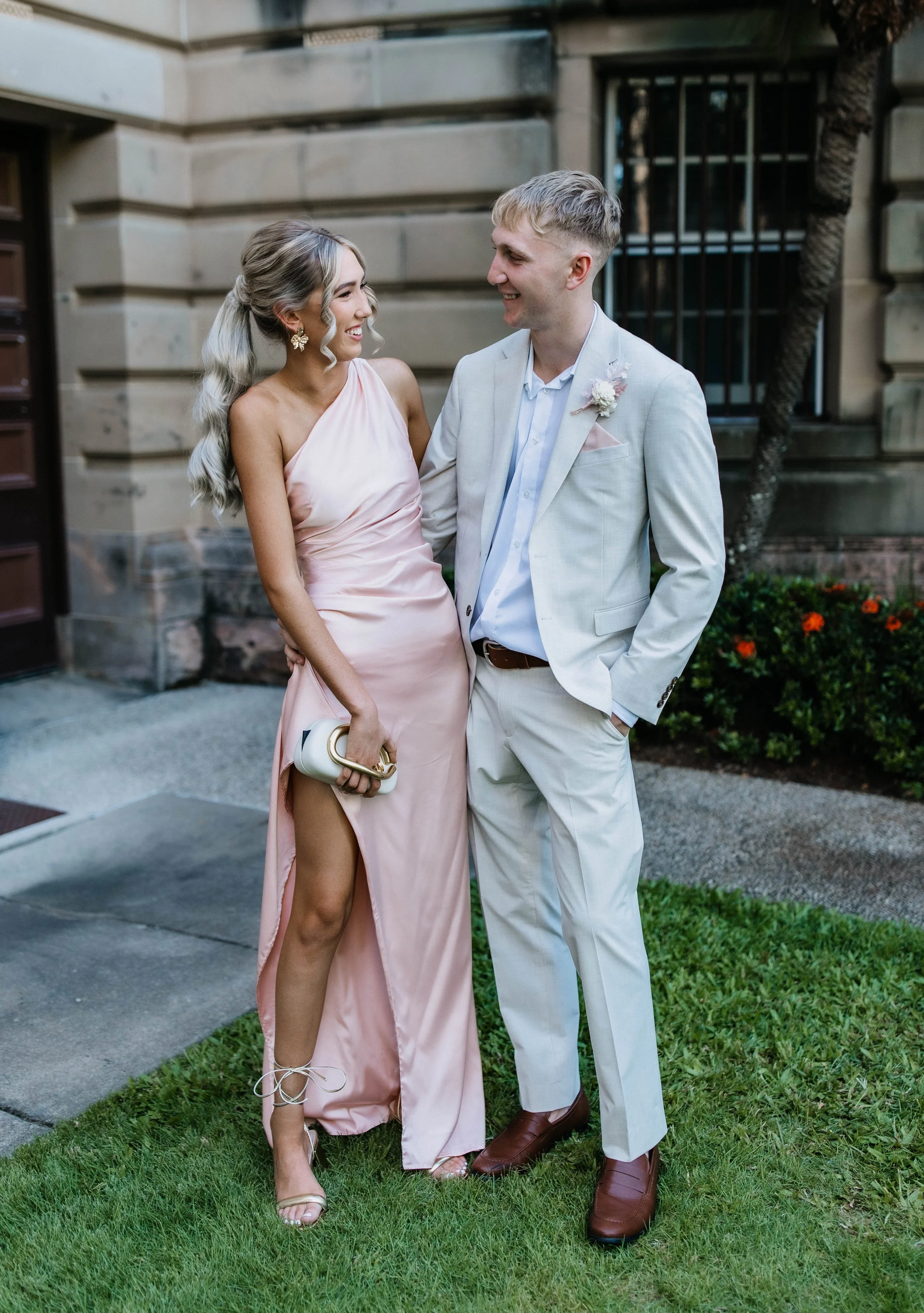 A young woman and a young man are standing outside, dressed formally and smiling at each other. The woman is wearing a light pink satin dress with a high slit, holding a small clutch, and has long, wavy gray hair. The man is wearing a light beige suit with a white shirt, brown shoes, and has short blond hair. They are standing on a grass lawn in front of a building with a window and a tree.