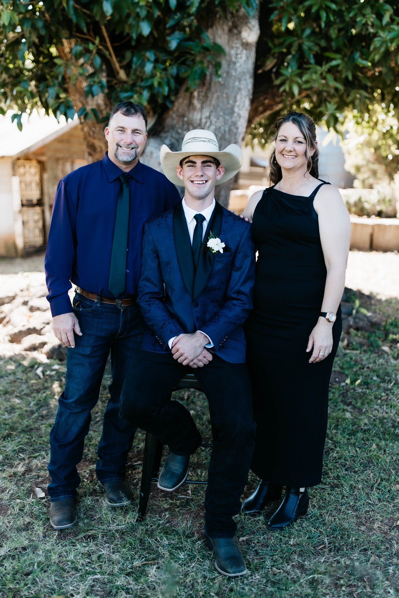 A family of three posing outdoors near a large tree. The father stands to the left, wearing a navy shirt and dark jeans. The mother stands to the right, wearing a black dress and black boots. The son, sitting in front, wears a dark suit with a boutonnière and a cowboy hat.