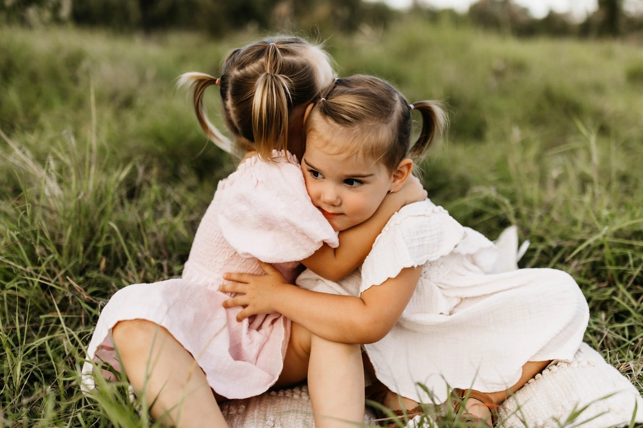 Two young girls with braided hair hugging each other while sitting in a grassy outdoor field.