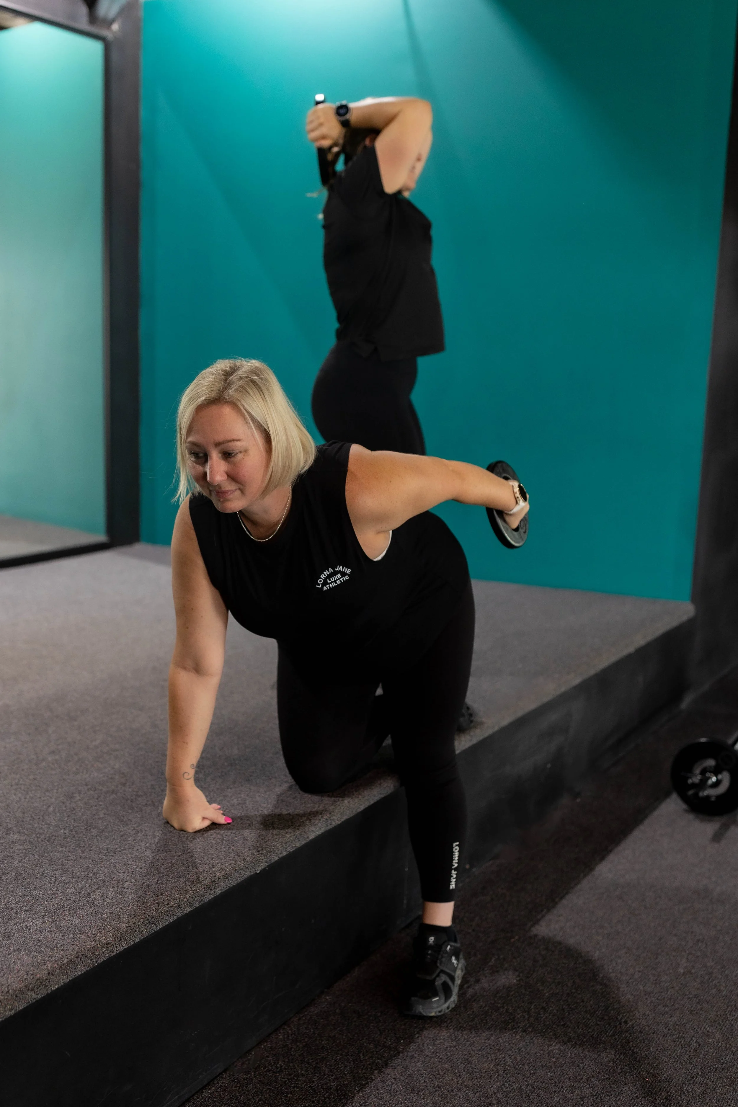 A blonde woman with a black athletic shirt and black leggings performing a workout exercise, supported on her right knee and left hand, in front of a gym mirror with a teal wall in the background.