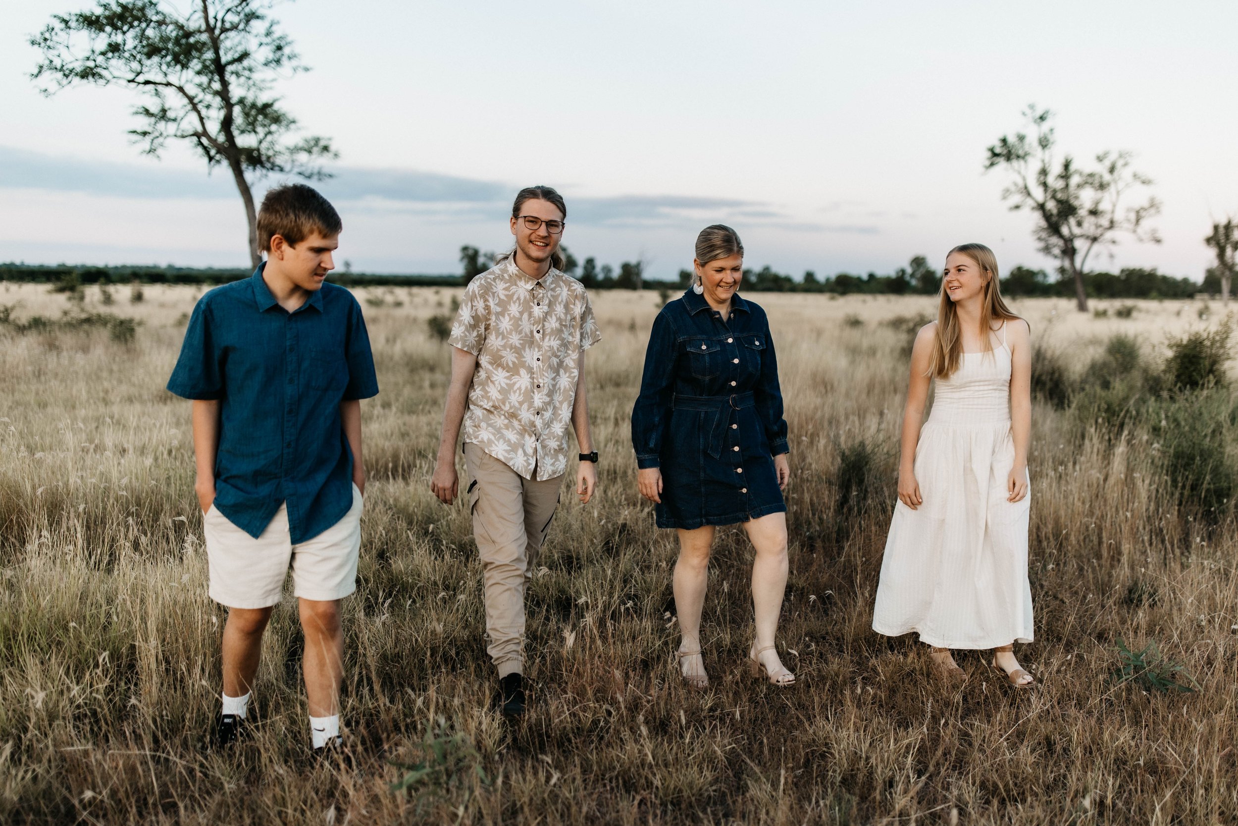 Four young adults walking through a grassy field with sparse trees under a cloudy sky, smiling and chatting.