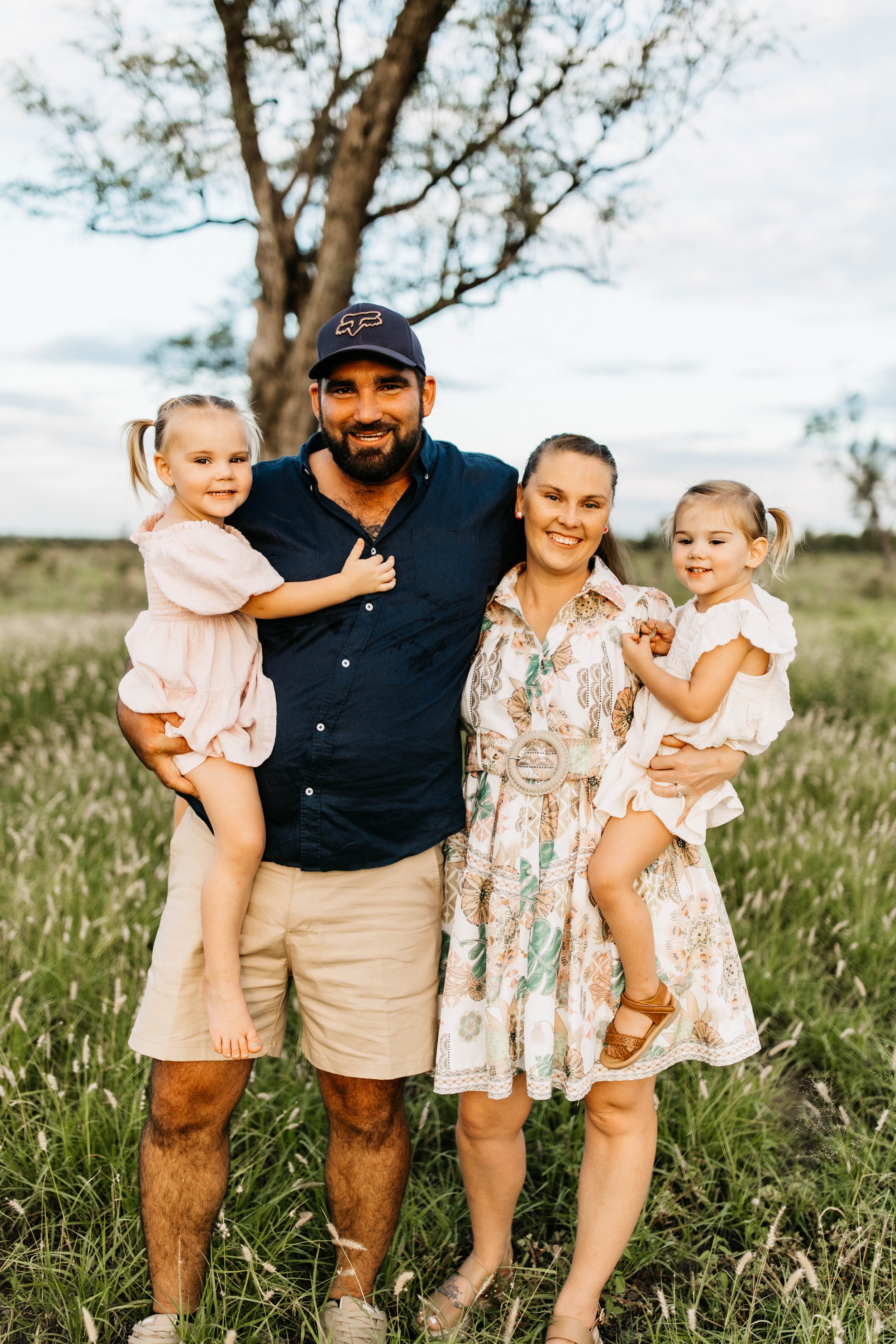 A happy family of four standing in a grassy field with a tree in the background. The father, on the left, has a beard, is wearing a navy cap, navy shirt, and khaki shorts. The mother, on the right, is holding two young girls, and is wearing a floral dress. The girls, both with pigtails, are dressed in light-colored dresses.