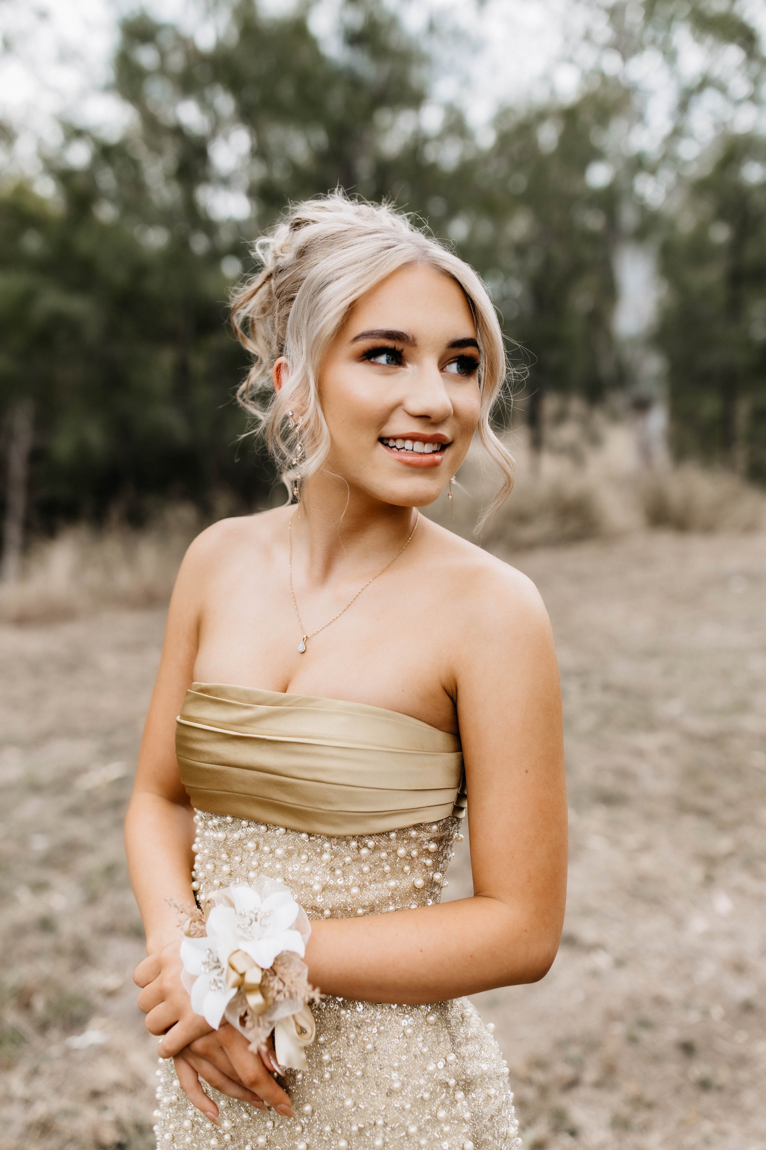 A young woman in an elegant champagne-colored dress with pearls, holding a small bouquet, standing outdoors in a natural setting with blurred trees in the background.