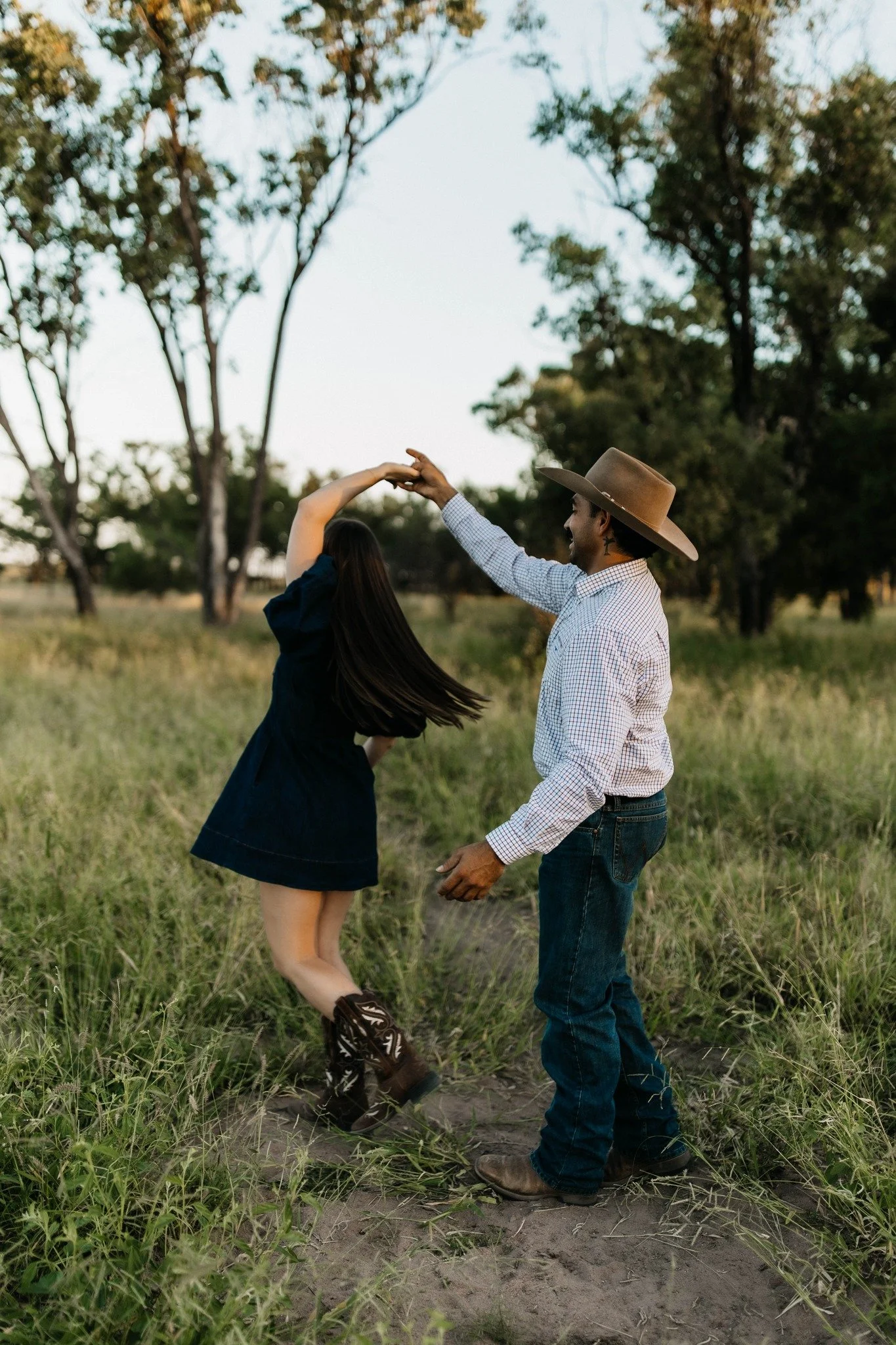 A man and woman dancing outdoors in a grassy field with trees in the background, as the sun sets. The woman wears a black dress and boots, while the man wears a cowboy hat, a checkered shirt, and jeans.