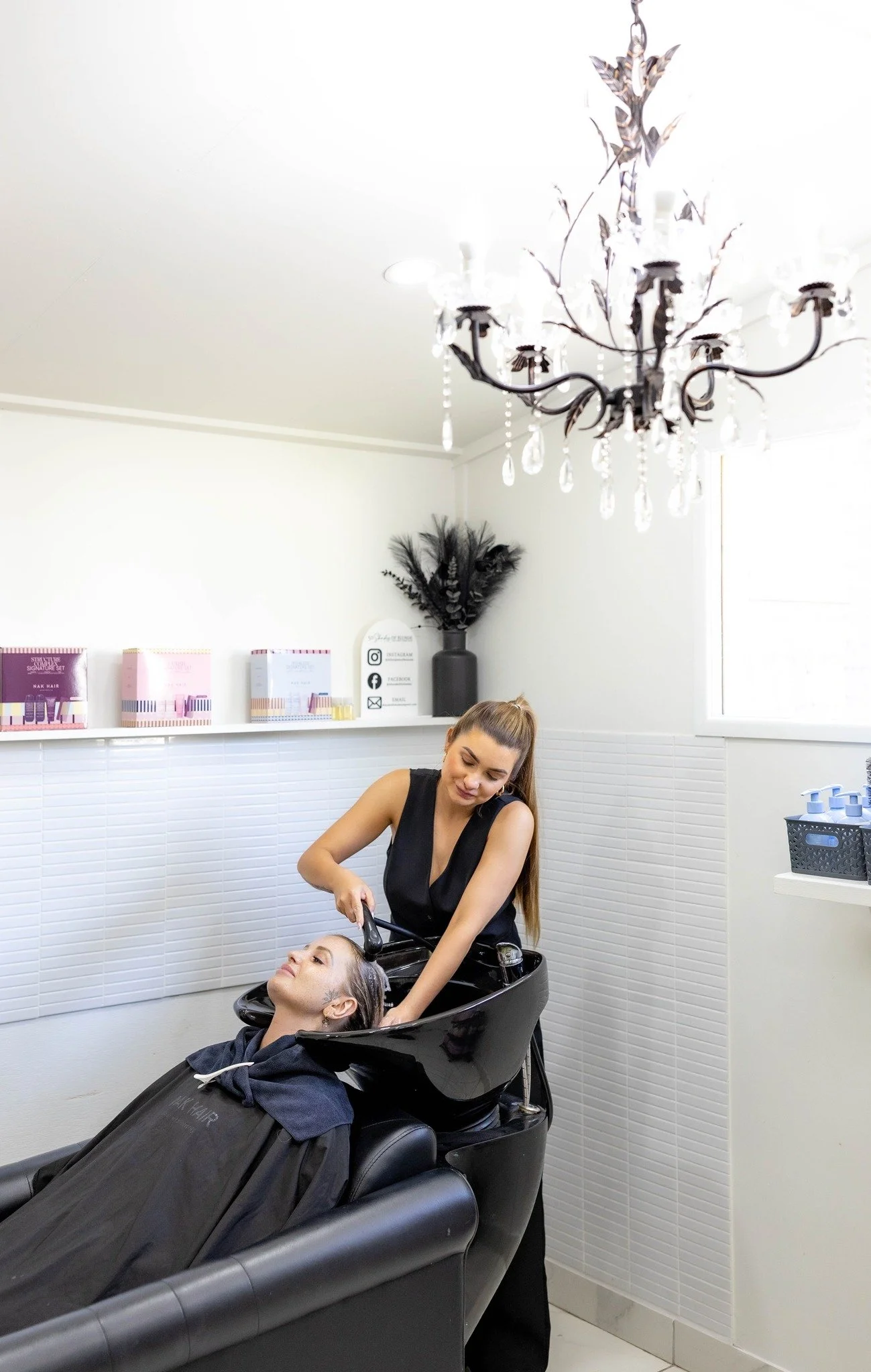A woman is sitting in a shampoo chair at a salon, getting her hair washed by a stylist. The stylist is standing behind her, washing her hair in a black sink. The salon has white walls, a chandelier hanging from the ceiling, and shelves with hair products.
