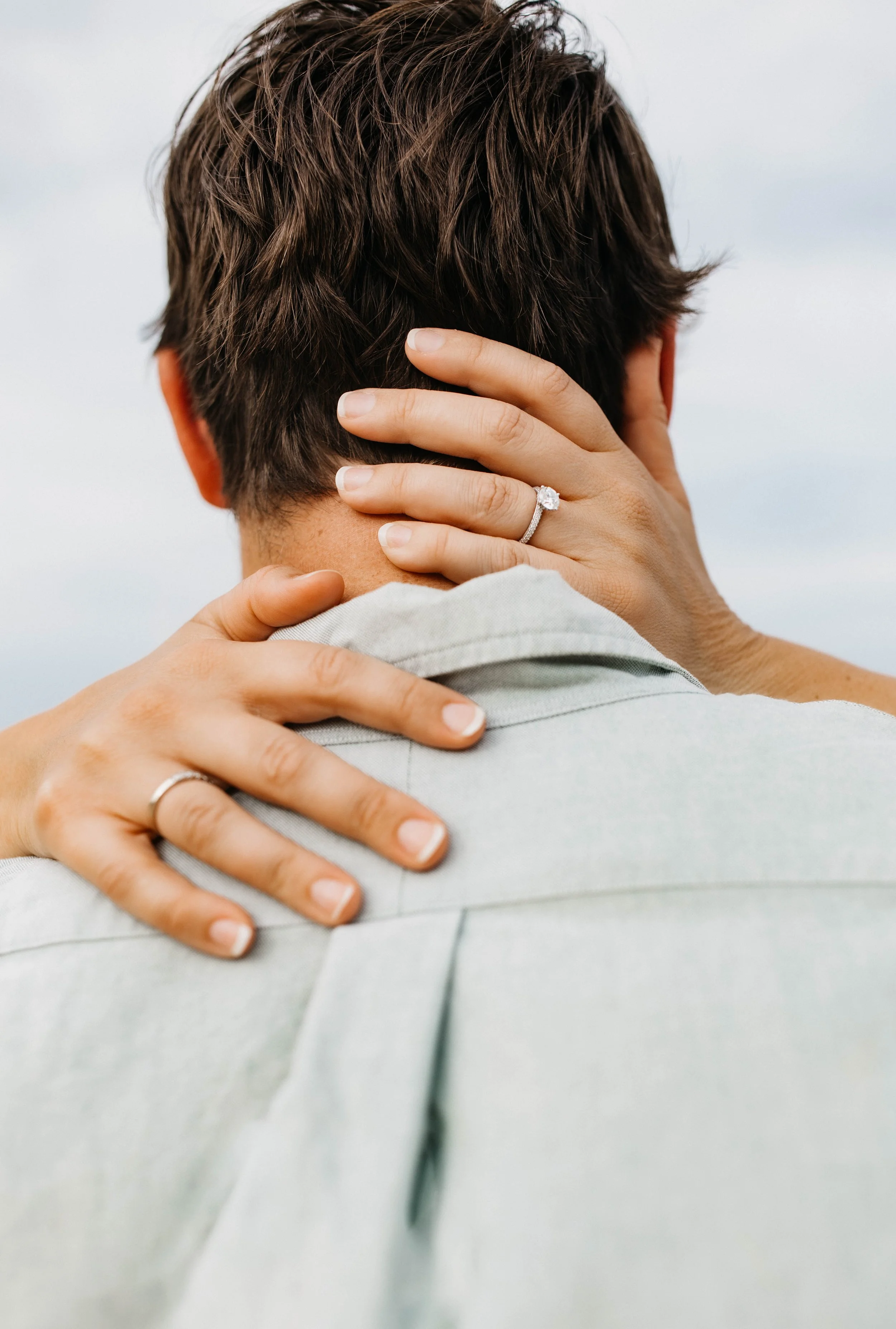 A person with short, dark hair is embracing another person, with their hand resting on the back of the other's head, showing an engagement ring. The background is blurred, and the focus is on the hands and hair.