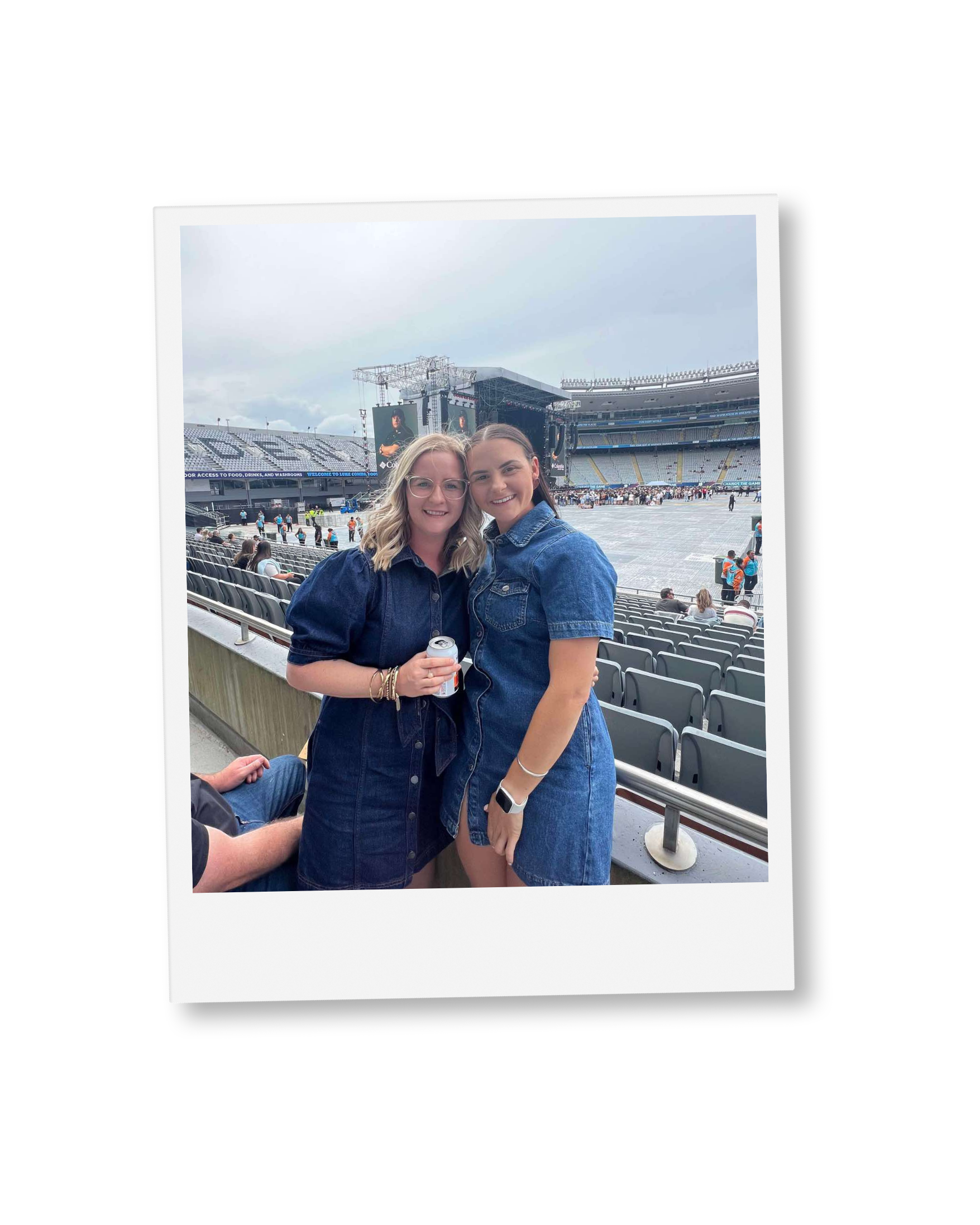 Two young women smiling and posing for a photo at an outdoor concert venue with a large stage, seating, and people in the background.
