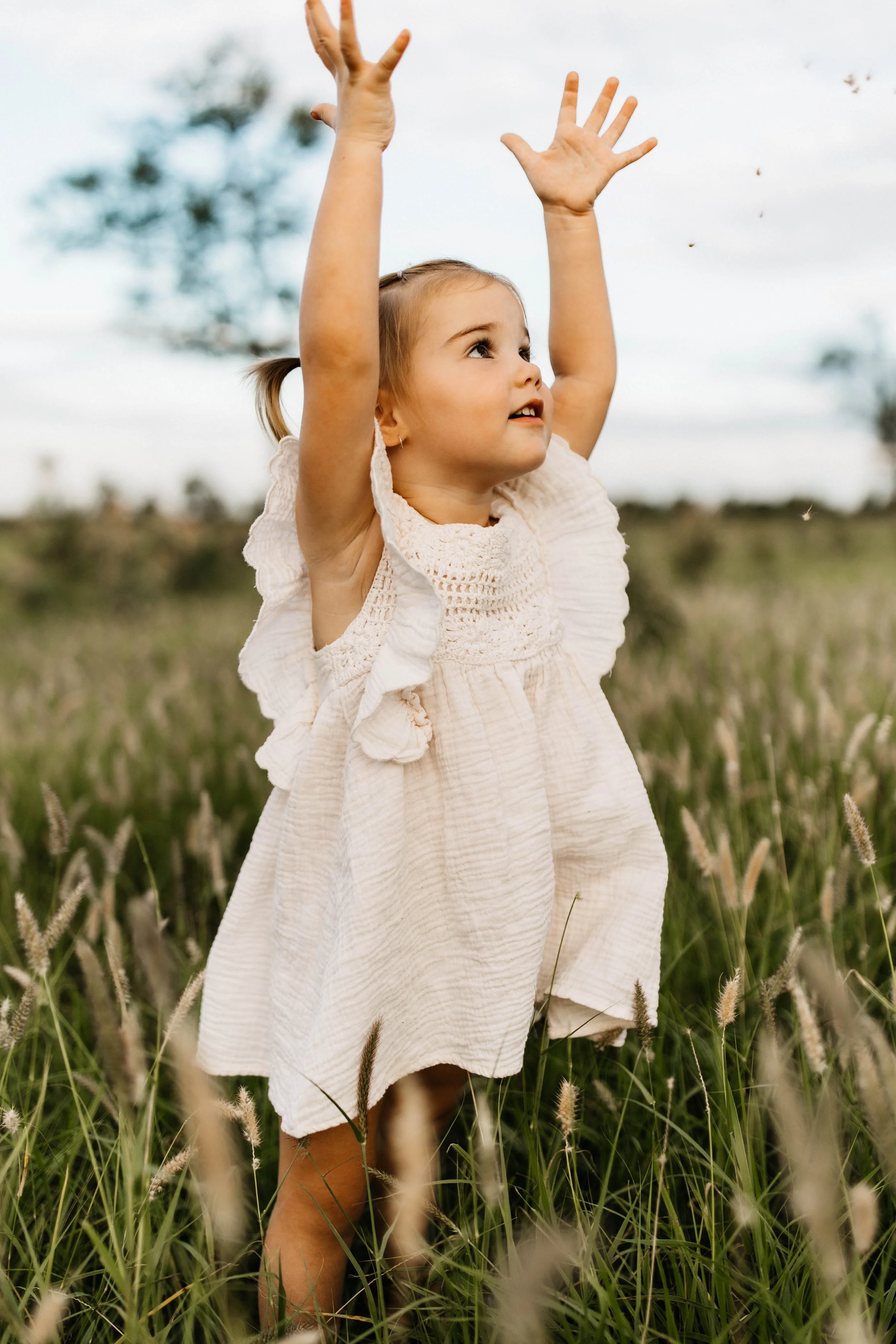 A young girl with pigtails wearing a beige dress standing in a field of tall grass, reaching up with both hands towards the sky, with two trees blurred in the background.