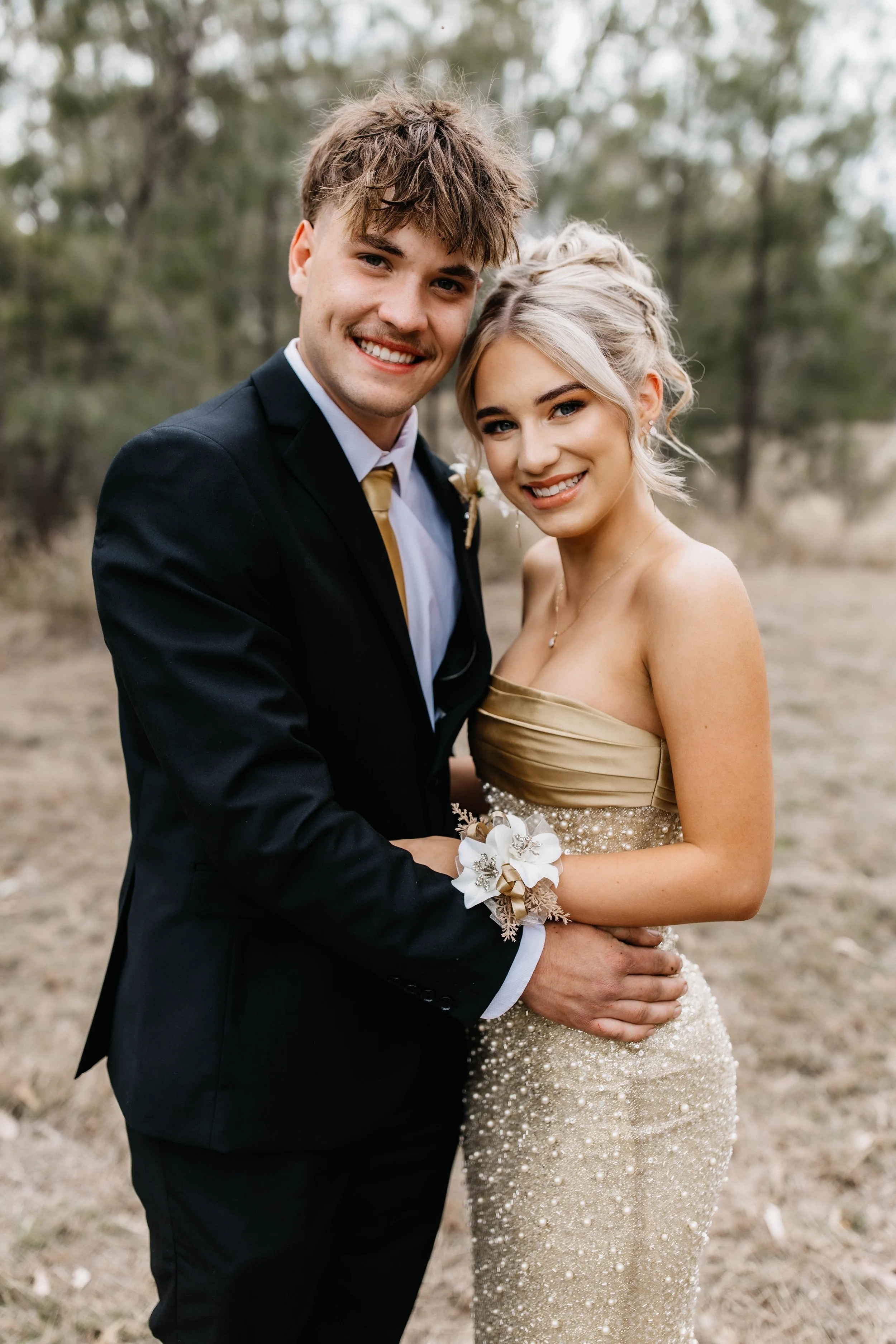 A newly married couple stands outdoors in a wooded area, smiling and holding each other. The groom wears a black suit and gold tie, and the bride wears a strapless, gold, sequined gown with a floral wrist corsage.