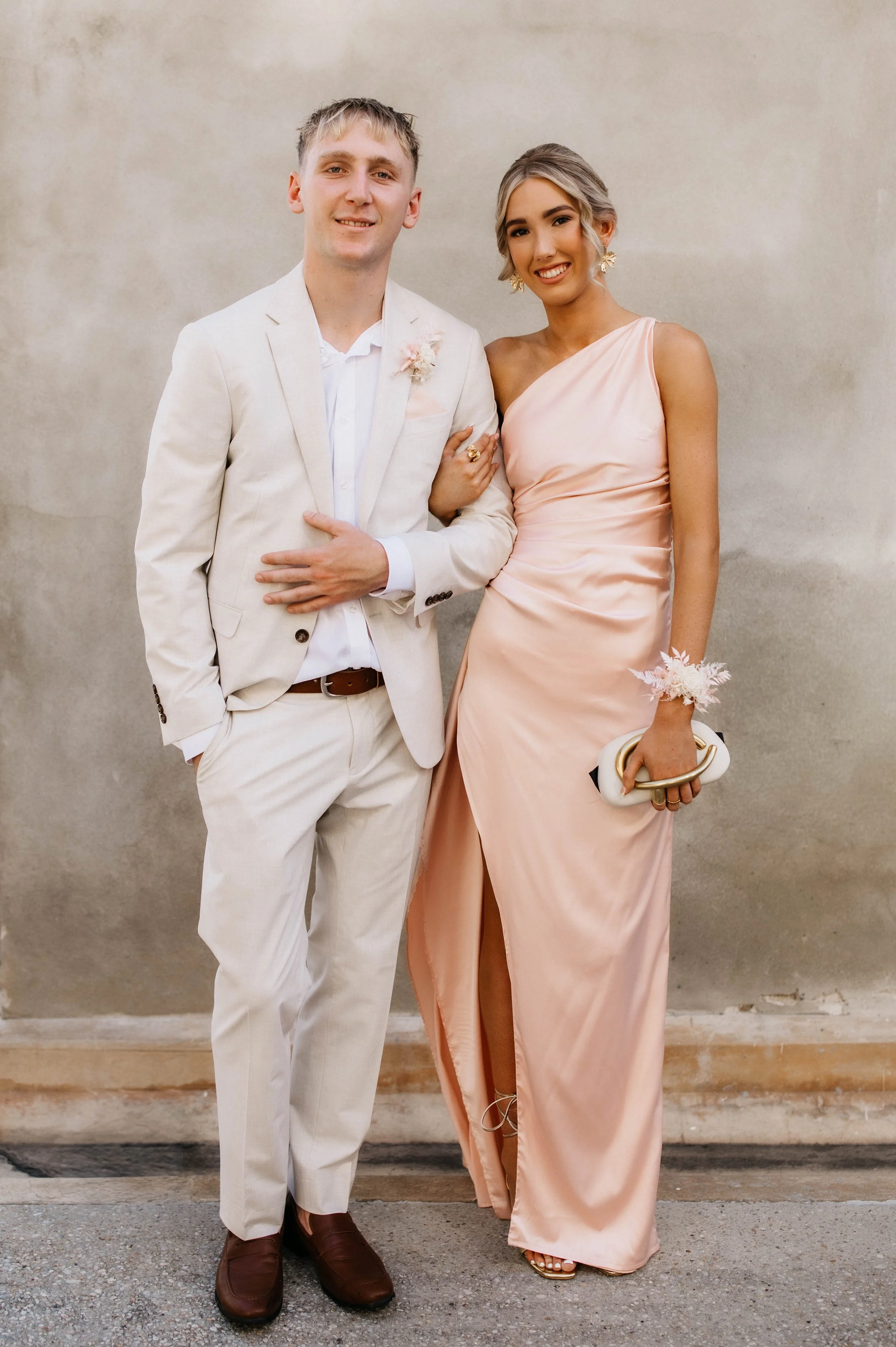 A man and woman dressed in formal attire standing side by side against a beige wall. The man is wearing a light-colored suit with a white shirt, and the woman is in a peach-colored satin dress with one shoulder strap. They are smiling and holding hands, with the woman's arm linked through the man's.