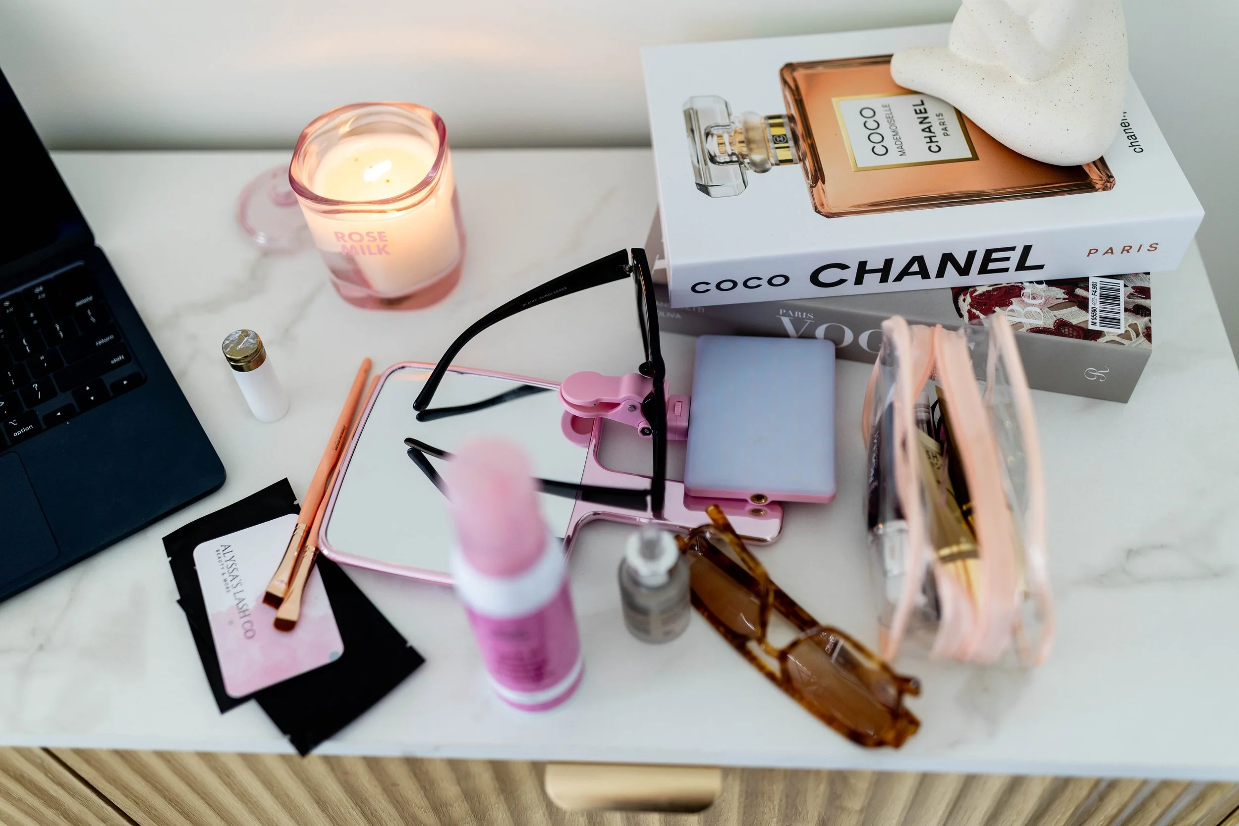 A cluttered desk with various beauty and fashion items, including a Chanel perfume box, sunglasses, a lipstick, skincare products, a candle labeled 'Rose Milk,' and a pack of mascara, along with a partially visible laptop.