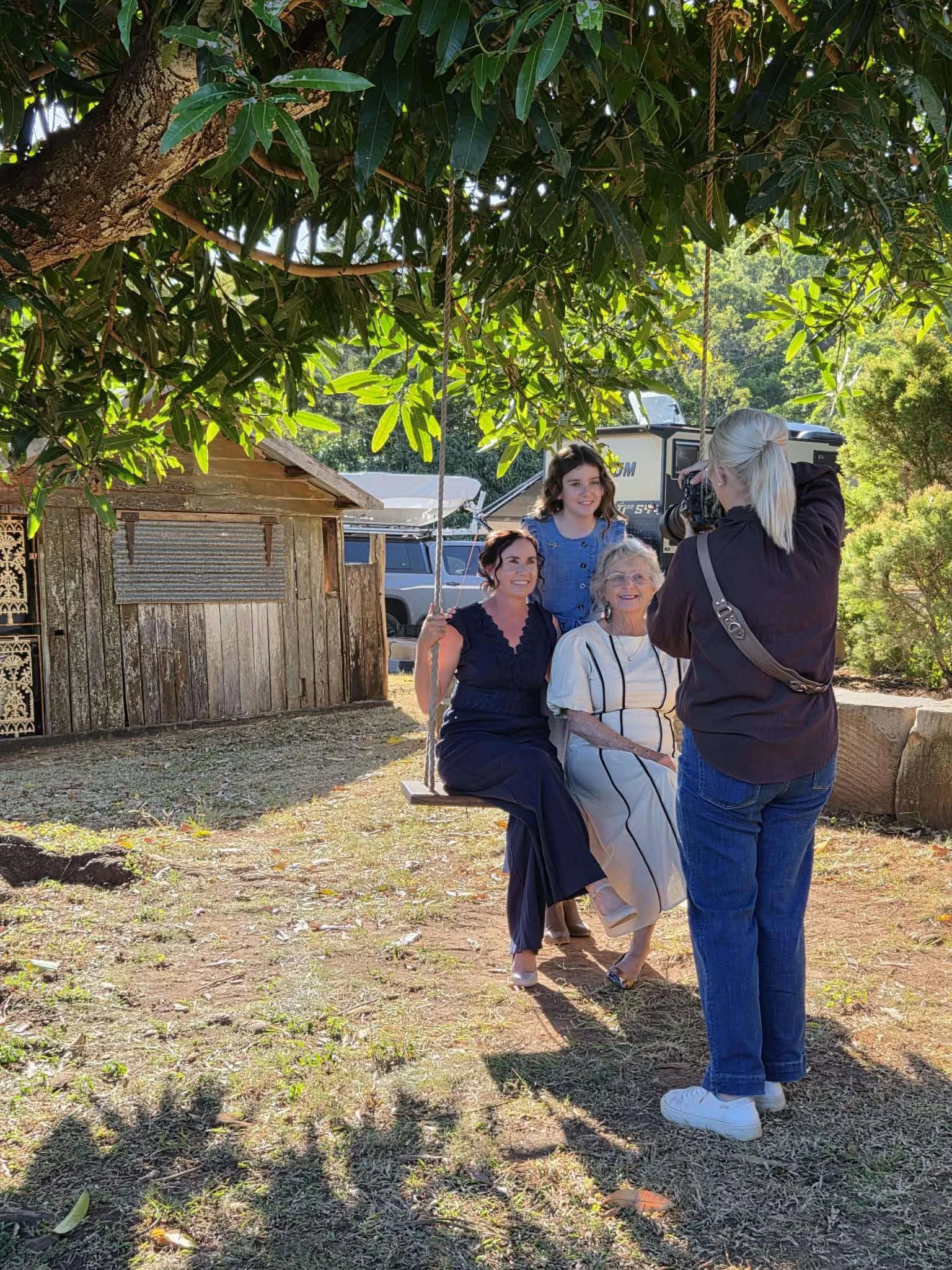 A woman is sitting on a swing held by a young girl, while another woman and an older woman stand nearby, being photographed outdoors under a large leafy tree during daylight. There is a rustic wooden shed in the background.
