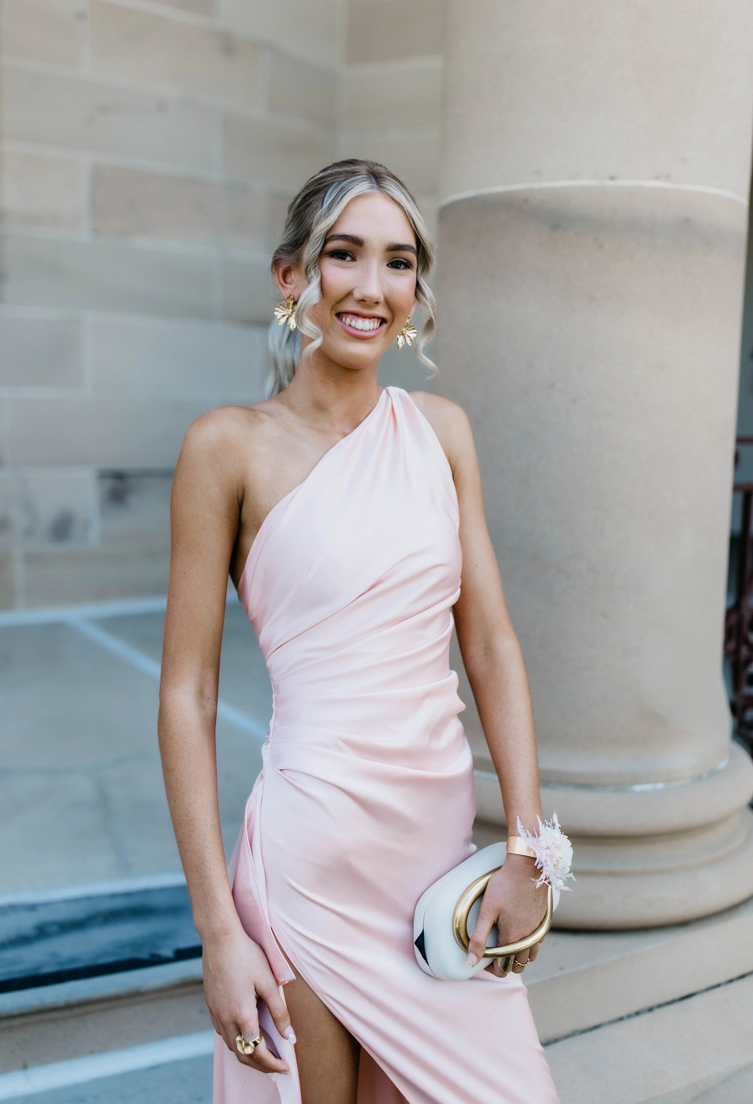 A woman in a light pink satin dress holding a white clutch with a gold handle, standing outdoors near a stone column and beige wall, smiling at the camera.