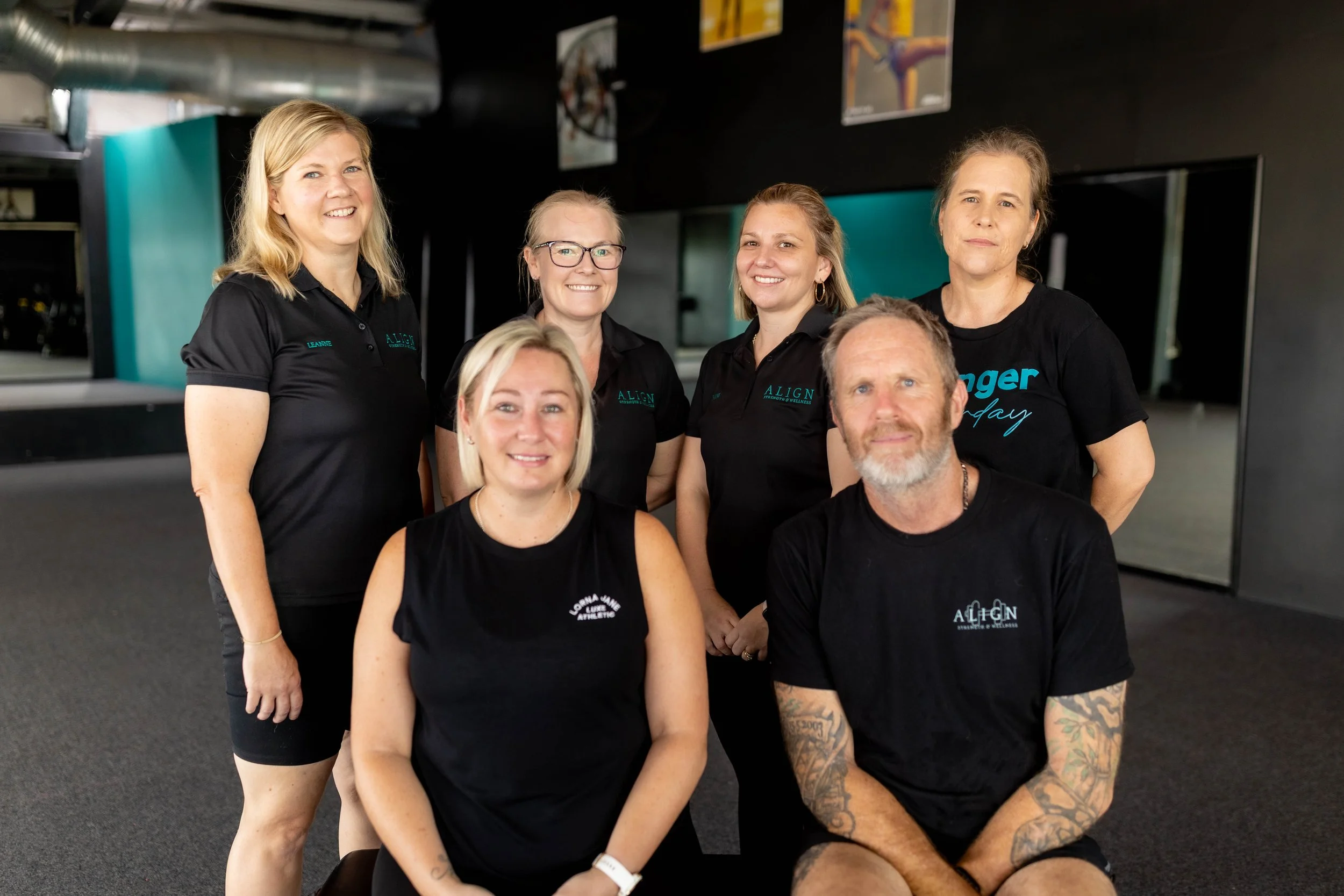 A group of six people, five women and one man, pose together indoors. They are dressed in black shirts, and some women are wearing glasses. The background includes a dark wall with colorful posters and a dark carpeted floor.